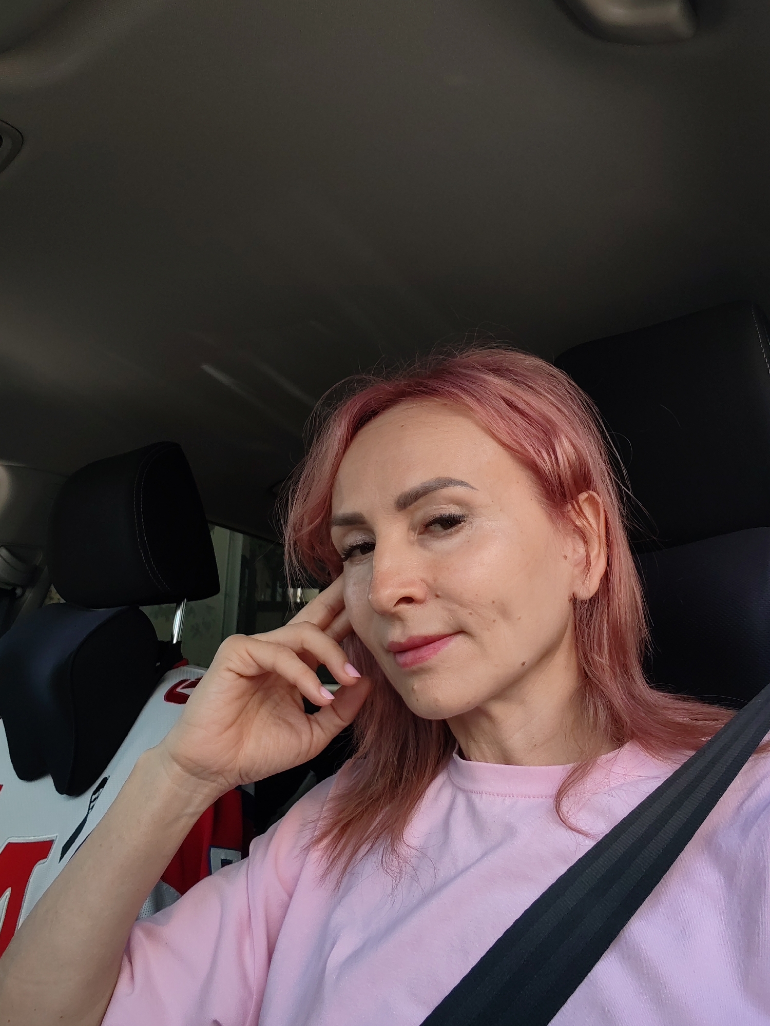 A woman with pink hair sits in the passenger seat of a car, her hand resting on her cheek. She wears a pink t-shirt and a black seatbelt is visible across her chest. The interior of the car is dark, with black headrests visible behind her. The image appears to be taken in Chaikovsky, Russia.