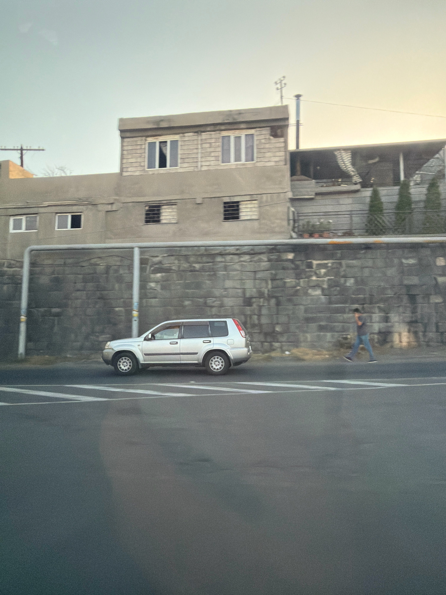 The image captures a street scene in Yerevan, Armenia, with a silver SUV driving on a road in the foreground. Behind the SUV, a pedestrian walks on a sidewalk next to a textured grey brick wall. A concrete building with multiple windows stands above the wall. The sky is a light blue, suggesting daytime.