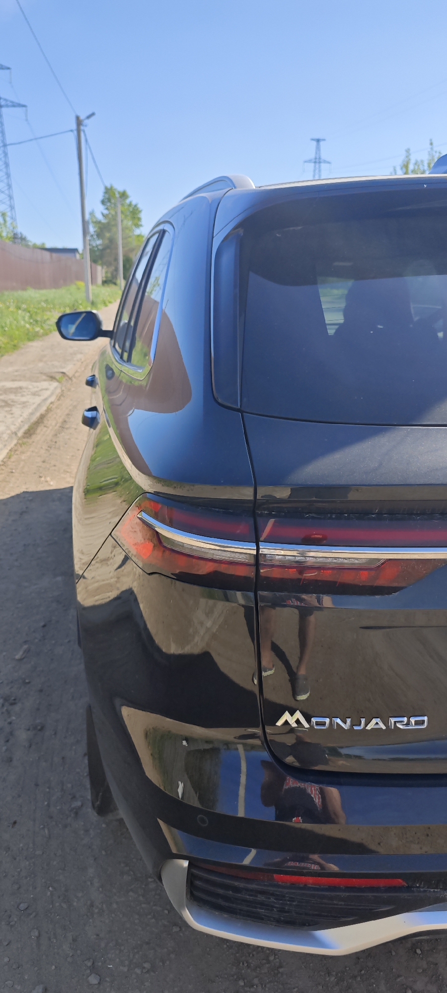 The image depicts the rear of a dark-colored car with the word "MONJARO" displayed in silver lettering. The vehicle is parked on a dirt road or shoulder. The reflection in the car's paint shows a person wearing shorts and shoes. The location is likely Chaikovsky, Russia, but this detail is not directly visible in the image. Power lines and poles are visible in the background, along with some vegetation and a fence.