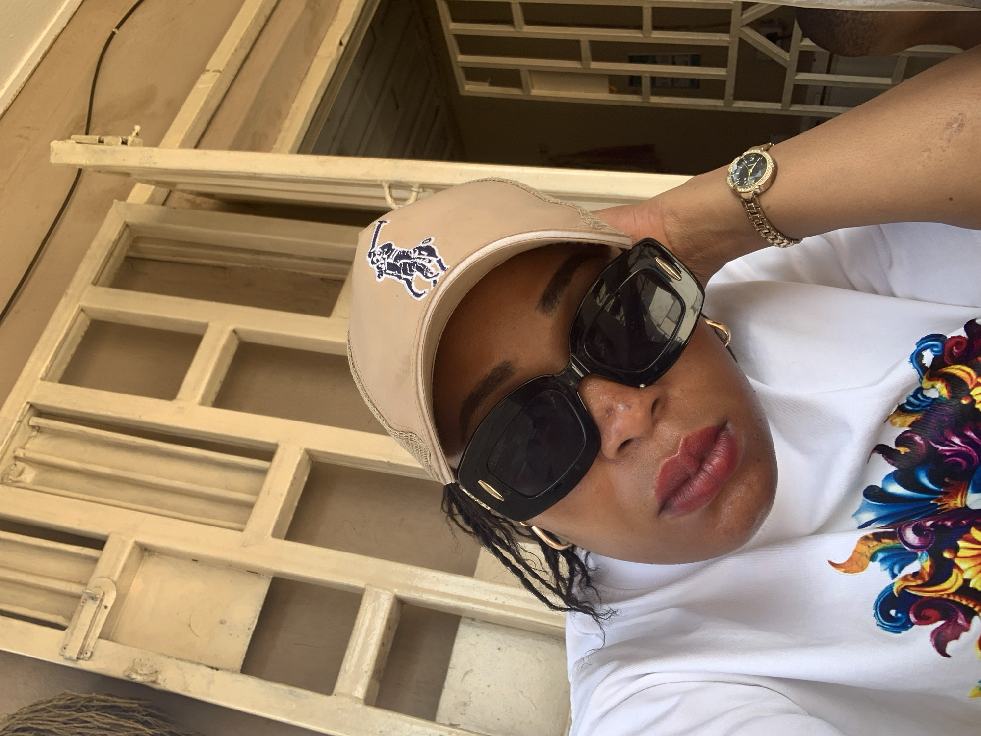 A medium shot taken in Sabon Tasha, Nigeria, features an African woman wearing a tan Ralph Lauren baseball cap, black sunglasses, and a gold wristwatch. She is wearing a white t-shirt with a vibrant, colorful floral design on the right side. She is positioned in front of a beige, segmented window frame.