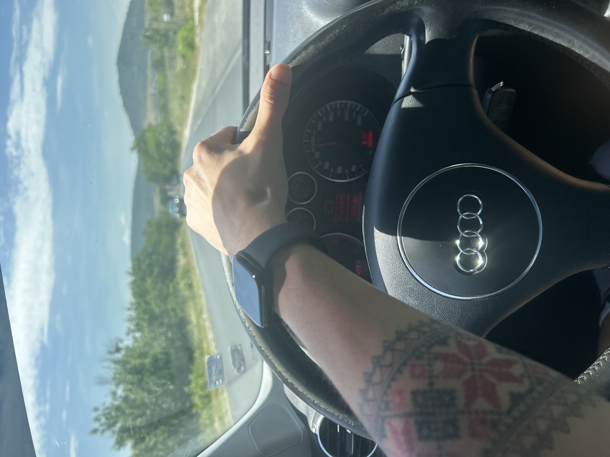 The image captures a first-person perspective from inside a car, showing a person's left arm and hand gripping a steering wheel while driving. The steering wheel prominently features the Audi logo, displaying four interlocked rings. On the dashboard, a speedometer with markings up to 260 is visible, alongside other gauges and a digital display showing "21.0 C" (likely temperature) and "0.0 km" on the trip meter. Several warning indicators are also present, including a red one. Car keys are visible in the ignition slot behind the steering wheel.
The person is wearing a black smart watch on their left wrist. A distinctive tattoo covers the forearm, featuring a folk-art-inspired pattern with red and grey/black geometric shapes, reminiscent of traditional Eastern European designs.
Outside the car, through the windshield, a sunny daytime landscape unfolds. The sky is bright blue with scattered white clouds. Rolling hills or mountains covered in green foliage are visible in the distance, alongside a road with visible markings and lush green trees closer to the car. The strong sunlight suggests mid-day, casting shadows and reflections on the windshield and car interior. The setting appears to be a rural or semi-rural area, consistent with the provided location context of Mihiltsi, Bulgaria.