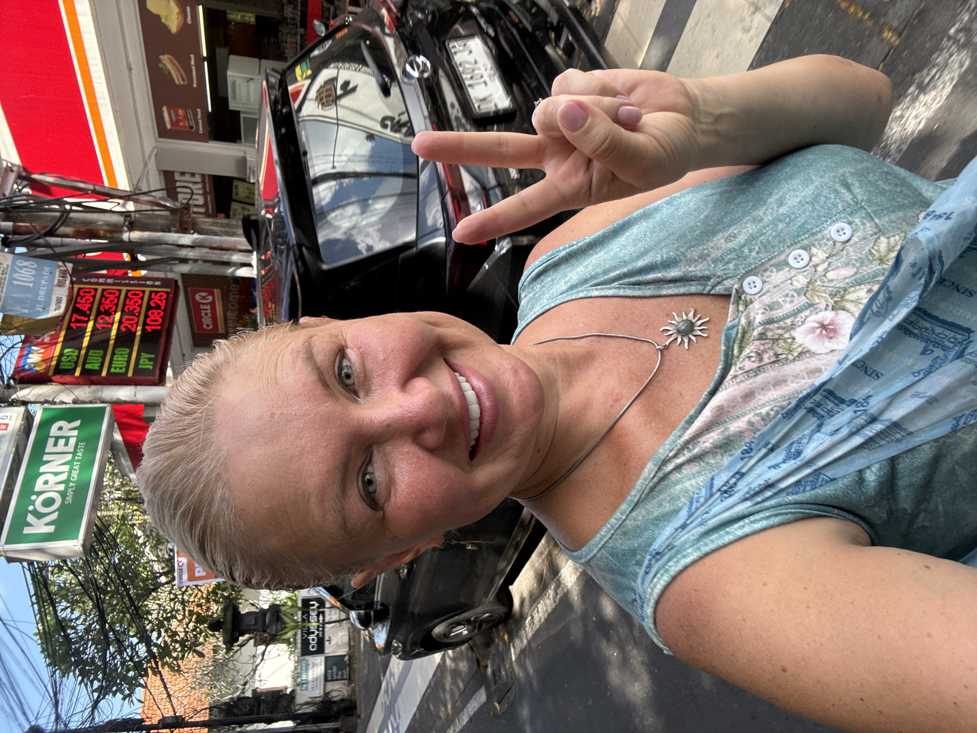 A woman is taking a selfie on a bright, sunny day on a street in Kuta, Indonesia. She has light hair and is smiling broadly, making a peace sign with her right hand. She is wearing a light blue sleeveless top with a floral pattern and a silver necklace with a distinctive sun-shaped pendant. Her fingernails are painted a light pink.
Behind the woman, a black car with a partial license plate "DK 1692 BJ" is visible, reflecting parts of the surrounding environment. The street scene is busy, characterized by numerous overhead electrical wires and commercial establishments. A red and white striped awning or building facade is prominent.
Several signs and texts are visible in the background. A green sign advertises "KÖRNER SIMPLY GREAT TASTE." A digital board displays currency exchange rates: "USD 17.450," "AUD 12.350," "EURO 20.350," and "JPY 108.25." Other signs include "JL DRUPADI 1068," a "CIRCLE K" logo, and advertisements for "VILLA ODYSSEY" and "VILLABANOOK," suggesting accommodation options. Storefront advertisements for "Processed Meat" and "Cheese" are also present. The bright lighting and clear shadows indicate it is daytime. The woman appears happy and is actively engaging with the camera, capturing her experience in this lively street setting.