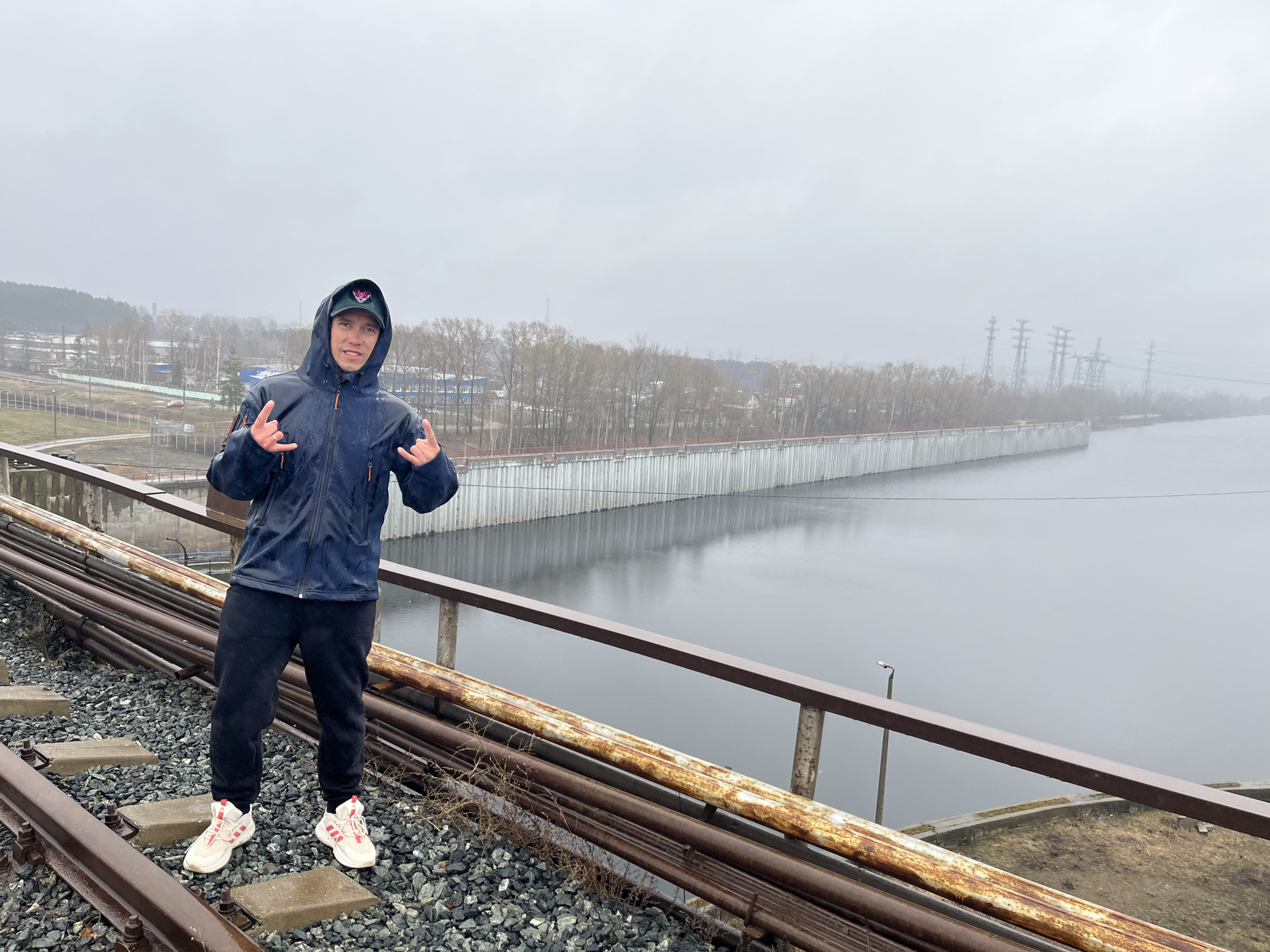 An outdoor, eye-level shot captures a man posing on railroad tracks next to a large body of water under a grey, overcast sky.
The primary subject is a young man, standing slightly left of center, facing the camera with a smile. He is wearing a dark blue hooded jacket with the hood up, black sweatpants, and white and red sneakers. His right hand is raised, and his left hand is held lower, both forming a "rock on" gesture with extended index and pinky fingers. He wears a dark baseball cap under his hood, with a distinctive pinkish-purple logo that appears to be a stylized "V" or similar emblem. The jacket appears wet, suggesting recent rain or damp conditions.
He stands on worn railroad tracks, with dark gravel filling the space between the ties. To his right, several rusty, thick pipes run parallel to the tracks and a rusty metal railing separates the tracks from the water. The water body, likely a river or reservoir, is a dark, still grey, reflecting the sky. Along the far bank of the water, a long, light-colored concrete retaining wall extends into the distance.
Beyond the retaining wall and water, a landscape of bare trees stretches across the horizon. In the far distance on the left, industrial-style buildings with grey roofs and fences are visible. Further to the right, several power line towers with complex lattice structures rise above the trees, partially obscured by the misty, grey atmosphere. The overall weather appears cold and damp, with a uniform, low-hanging overcast sky.
The scene depicts an industrial or semi-industrial setting, specifically in Chaikovsky, Russia, characterized by infrastructure like railroad tracks, pipes, and power lines adjacent to a natural water feature.