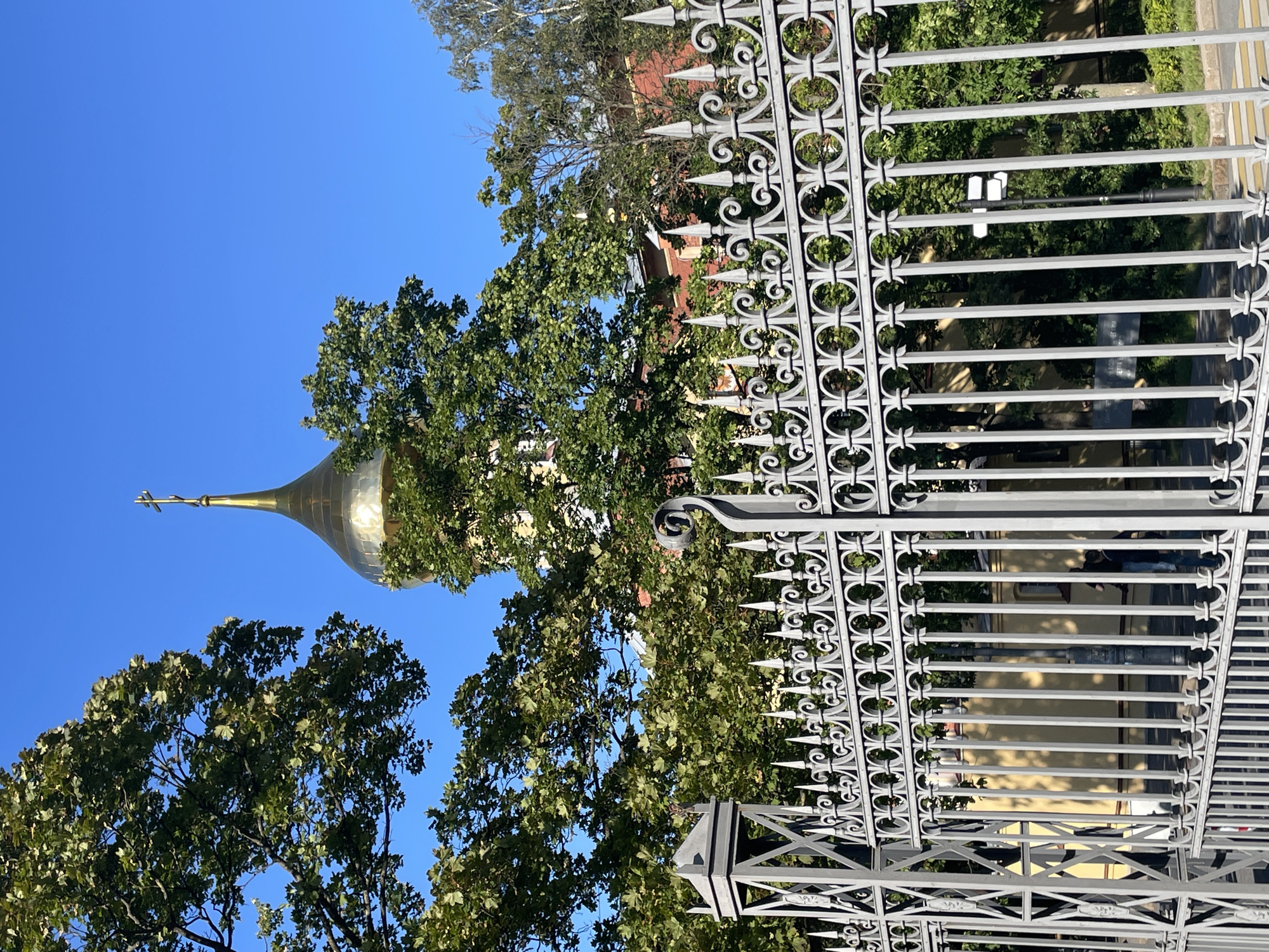 A sunny day is captured in an outdoor shot of a golden dome of a church or cathedral peeking through lush green trees. In the foreground, a decorative grey metal fence with pointed finials and circular motifs spans the width of the frame. The fence is intricate and appears to be a boundary to an estate or a public space. Behind the fence, the trees are in full leaf, suggesting a warm season like summer or early autumn. The dome is prominently visible, topped with a cross, and reflects the bright sunlight. The sky is a clear, deep blue. The scene is likely located in Saint Petersburg, Russia, given the architectural style of the dome and the presence of mature trees often found in the city's parks and historical areas.