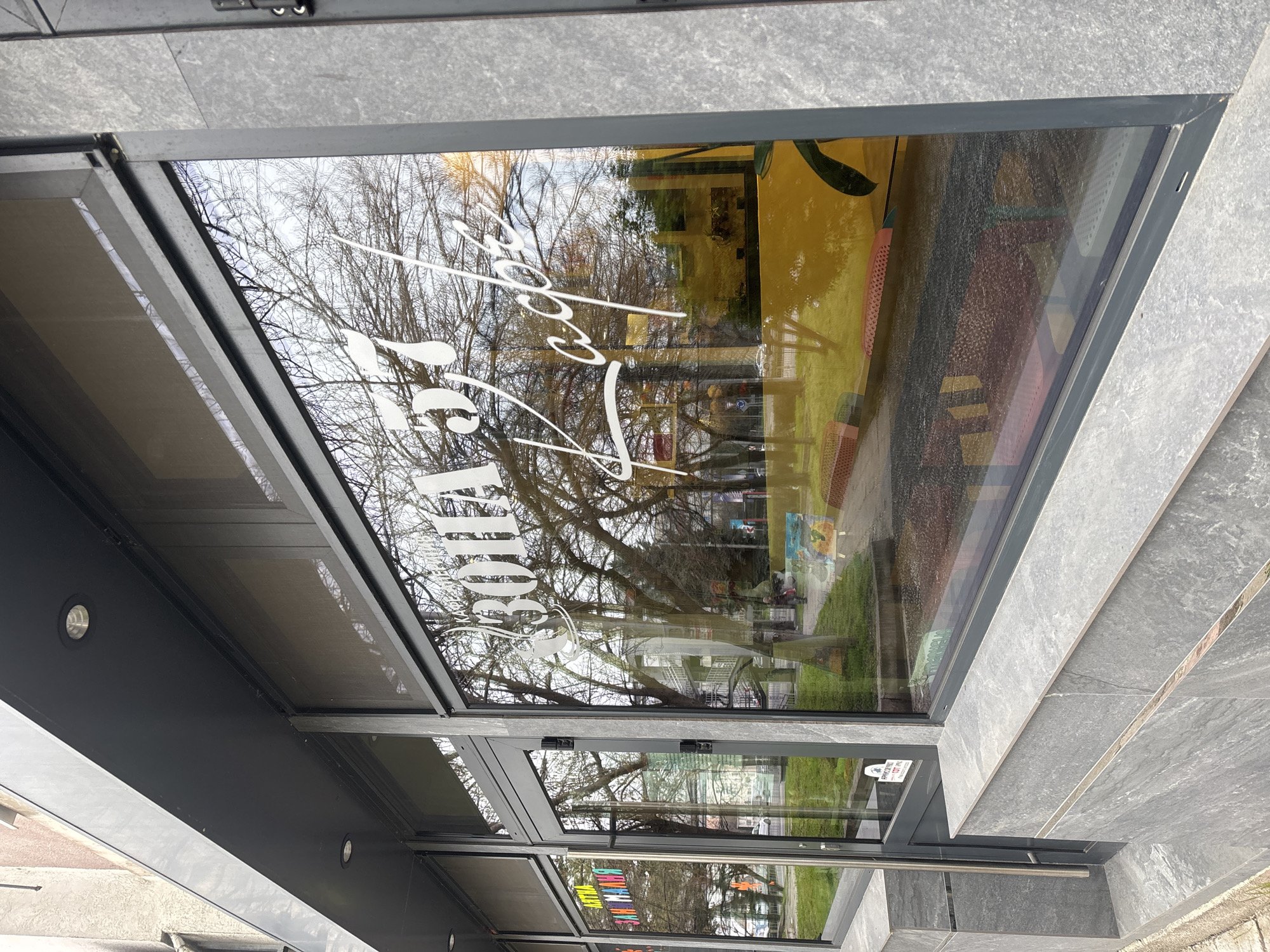 The image shows the exterior of a building, most likely a commercial establishment, in Burgas, Bulgaria. A large window dominates the view, reflecting the exterior environment – bare trees against a cloudy sky, a park or green space, and possibly other buildings. The window has white lettering on it (likely advertising). The building has a modern architectural style with grey stone or tile facade. A metal frame surrounds the window, with the building’s entrance or further windows visible beneath. The overall scene appears to be a public area, perhaps a street or park facing the building.