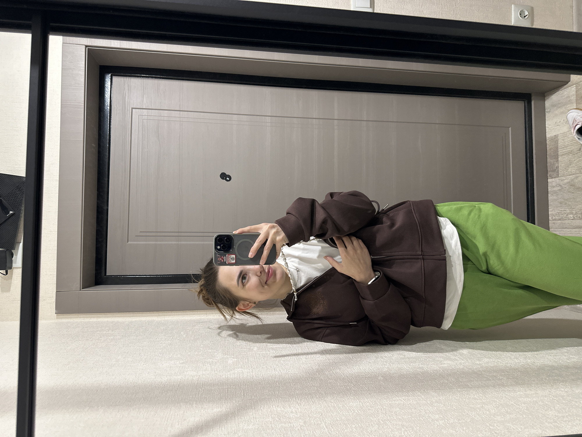 A young woman captures a mirror selfie in an indoor setting. She stands on a light-colored carpet or rug, facing a mirror that reflects her and the room behind her. She is casually dressed in a brown zip-up hoodie worn over a white t-shirt, paired with bright lime green sweatpants. A pearl necklace is visible around her neck. She holds a dark-colored smartphone, likely an iPhone with a multi-camera system and a magnetic ring on the back, up to take the photo, and she has a faint, relaxed smile.
Behind her, a light beige door with a prominent dark frame is visible. On the door, there's a small, dark, circular object, possibly a peephole. Above the door frame, on the light-colored wall, a silver electrical outlet is present. To the far right of the image, part of a light-colored sneaker is visible. The scene is brightly lit, suggesting daytime or a well-lit interior. The overall atmosphere is casual and relaxed. The photograph is taken in Perm, Russia. No discernible text is visible.