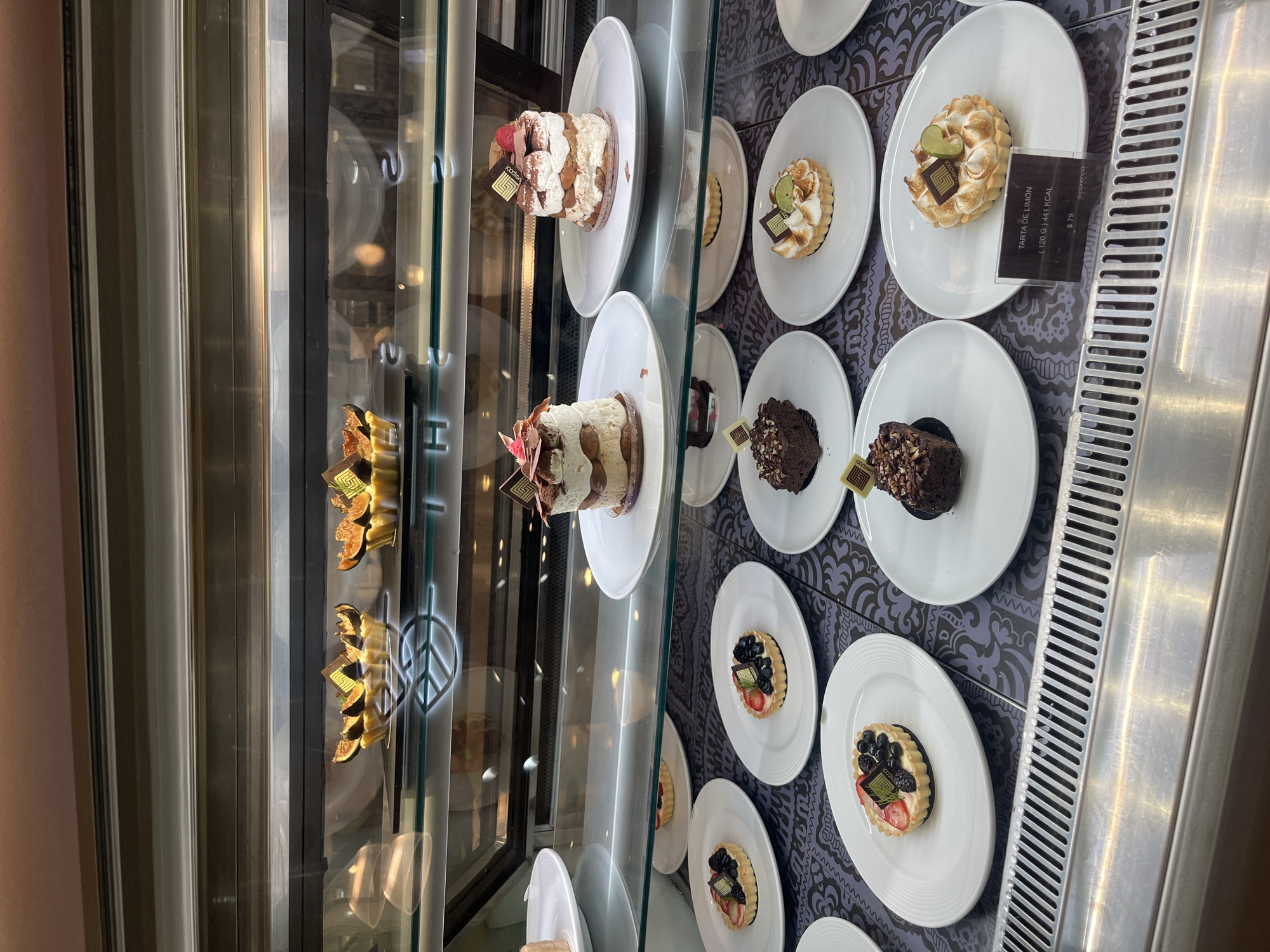 A refrigerated display case contains an assortment of individual cakes and tarts, each presented on a white plate. The items are arranged in vertical rows, suggesting multiple shelves within the case. The display case's interior backdrop is a patterned surface. Some items have price tags visible, indicating the cost of each dessert. The overall setting appears to be a bakery or dessert shop in Puebla, Mexico, indicated by the "Godiva" branding on some cakes.