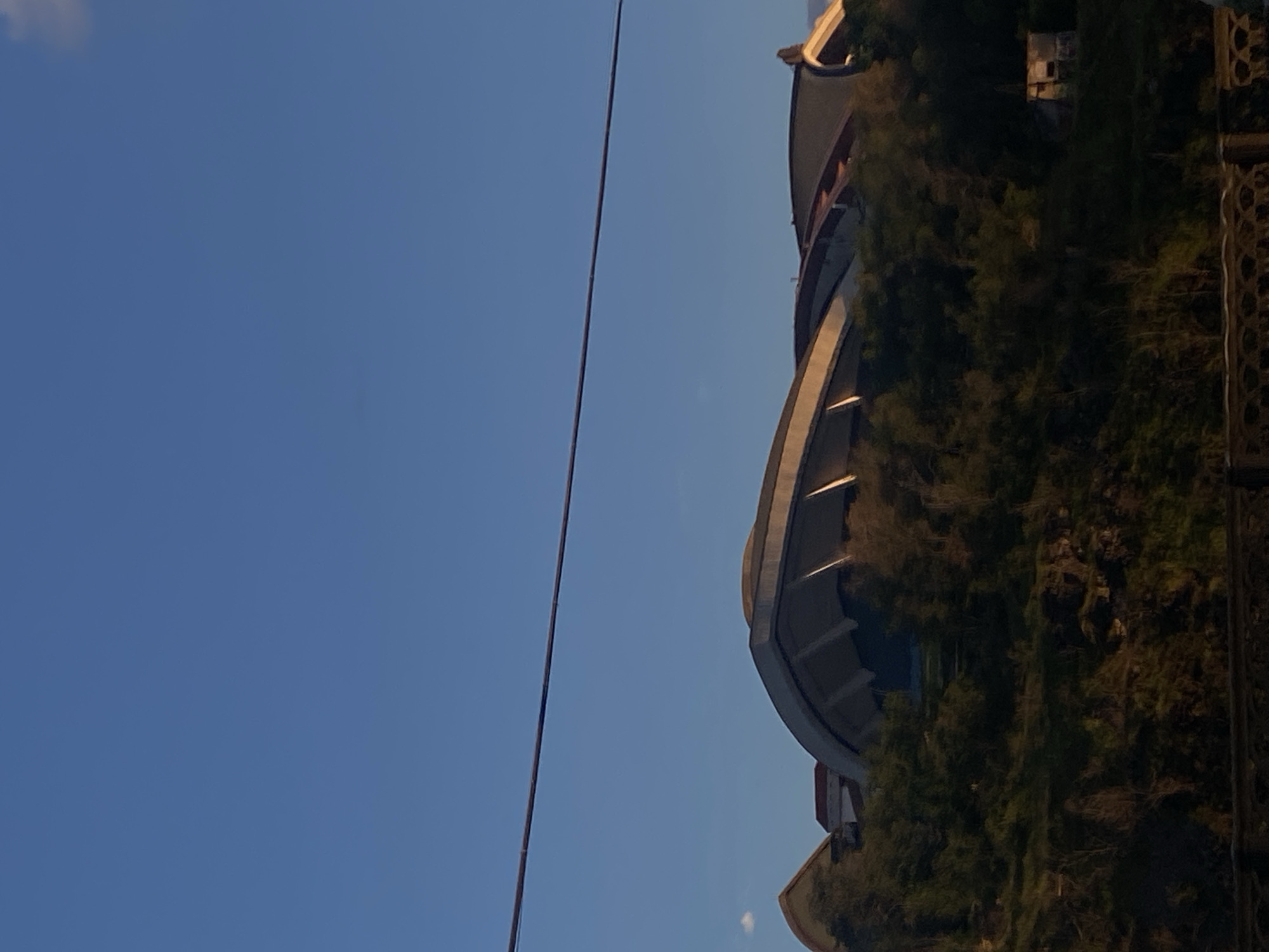 The image shows a partial view of a building, possibly a stadium or large structure, heavily integrated into a rocky, overgrown hillside. The structure features curved sections with some visible tiers or levels. Dense vegetation, including trees and other plants, covers a large portion of the hillside around the building. A thin, dark cable extends vertically across much of the left side of the frame, contrasting against a clear, blue sky. To the right of the building, a decorative, patterned metal structure appears. The image is taken at an upward angle, emphasizing the height of the building and its integration with the natural environment.