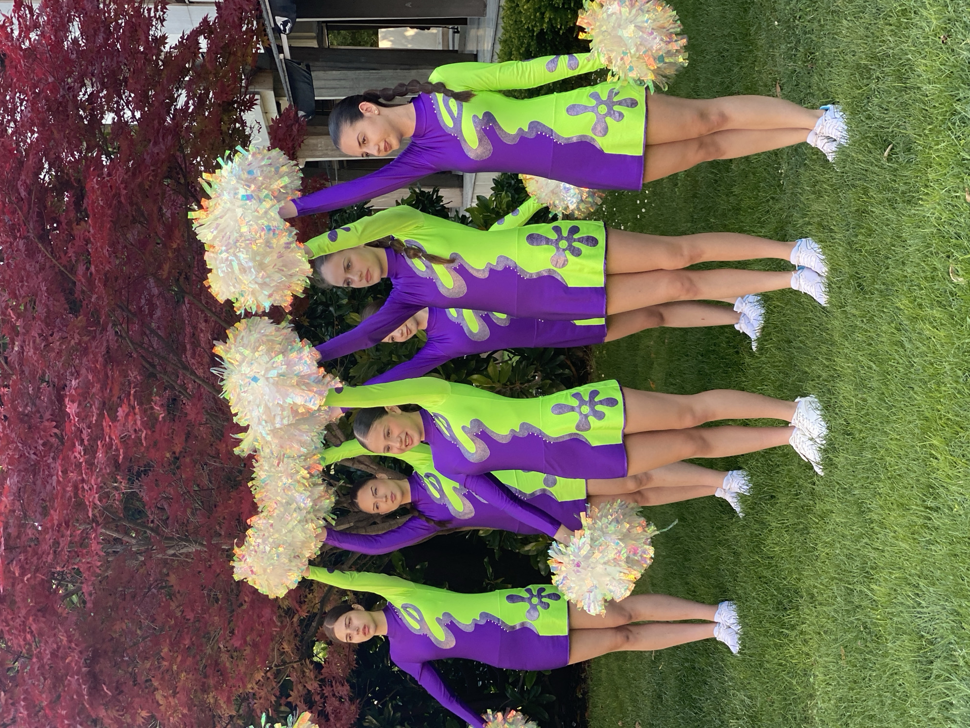 Six young females, appearing to be in their mid to late teens, are positioned on a grassy area, likely a lawn. They are dressed in matching cheerleader uniforms consisting of purple and neon green dresses with floral appliqué designs. Each individual is holding a large, shimmering pom-pom, with some pom-poms overlapping and extending out of the frame. The subjects are arranged in a staggered formation, facing forward. Behind them, a vibrant red-leafed tree dominates the background, with some foliage and architectural structures, including a building with windows, visible to the right. The lighting suggests an outdoor setting during daylight hours, possibly late afternoon due to the warm tones. The location is identified as Pleven, Bulgaria.