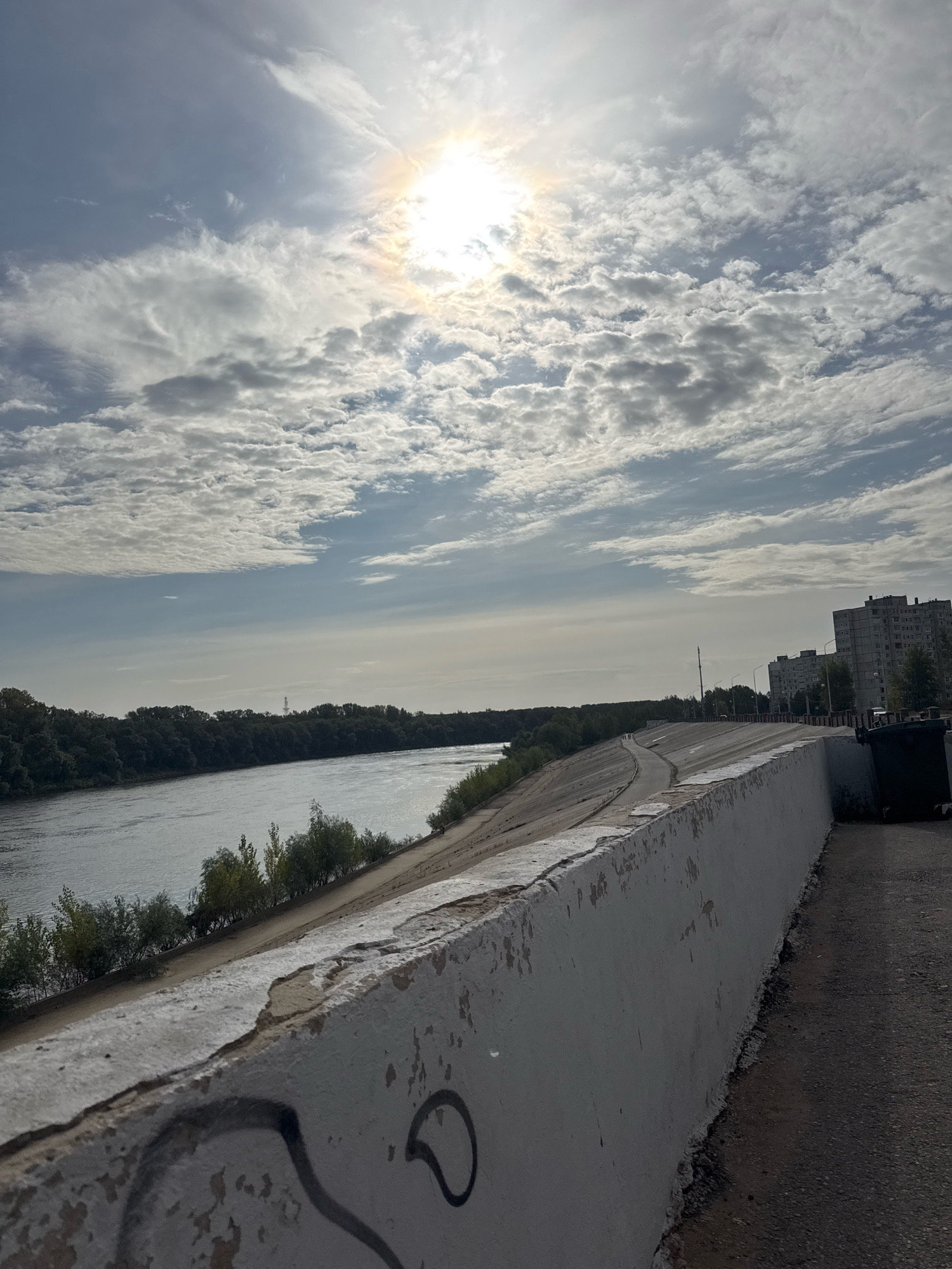The image shows a wide river on the left, bordered by a tree-lined bank. A concrete embankment runs along the right side of the river, with a paved path on top. The embankment wall is weathered and shows signs of graffiti. In the distance, on the right side of the image, are several tall apartment buildings. The sky is partly cloudy with the sun visible, creating a bright halo effect. The scene appears to be a waterfront area in a city, possibly Ufa, Russia, given the architectural style of the buildings and the presence of a large river.