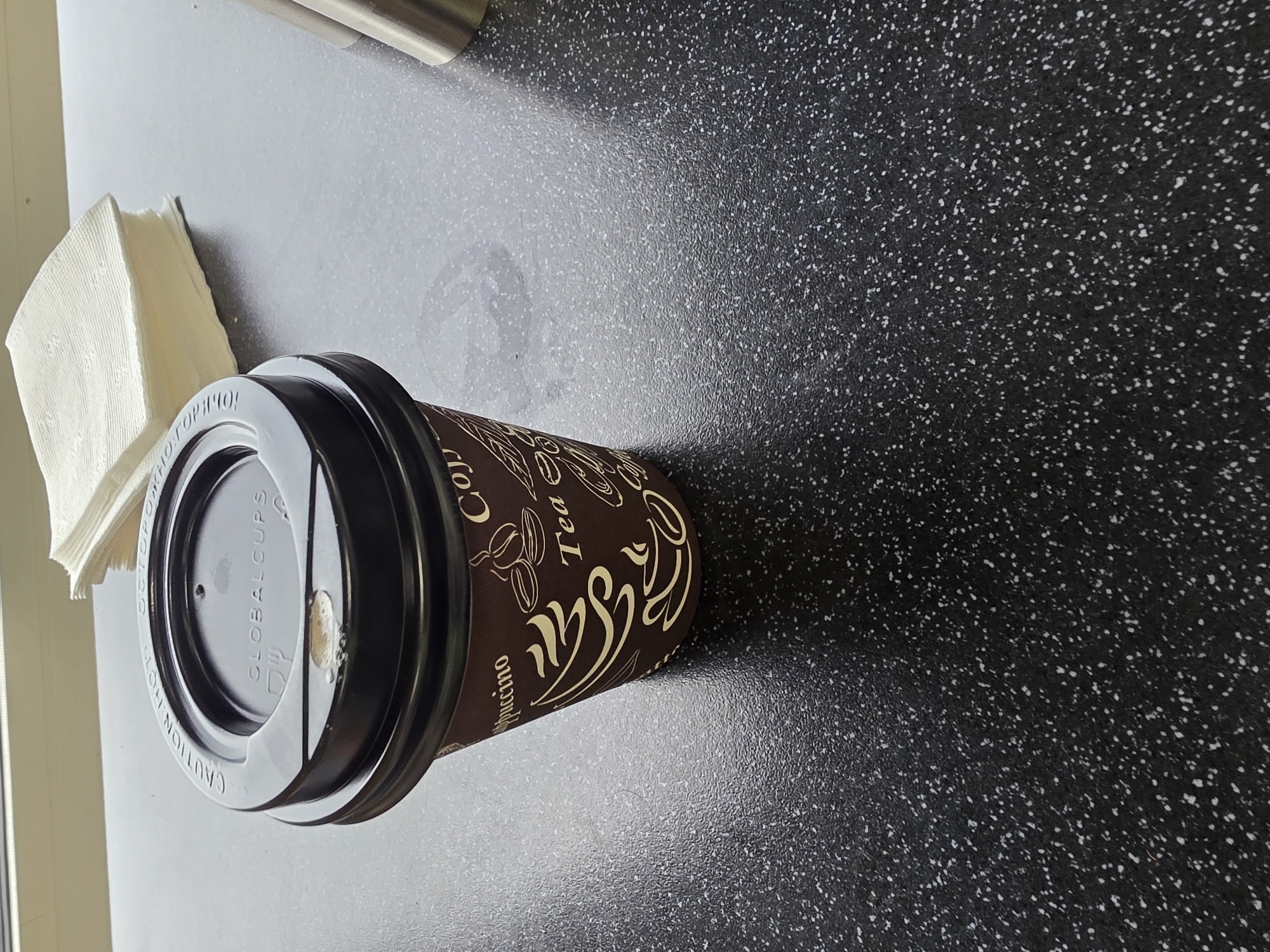 A coffee cup with a black lid sits on a dark speckled countertop. The cup has "Globalcups" printed on the lid and a brown paper sleeve with a pattern of coffee beans and cups. A stack of white napkins is placed behind the cup. A metal object, possibly a creamer dispenser, is partially visible to the right of the cup. The scene appears to be indoors, possibly in a cafe or office break room.