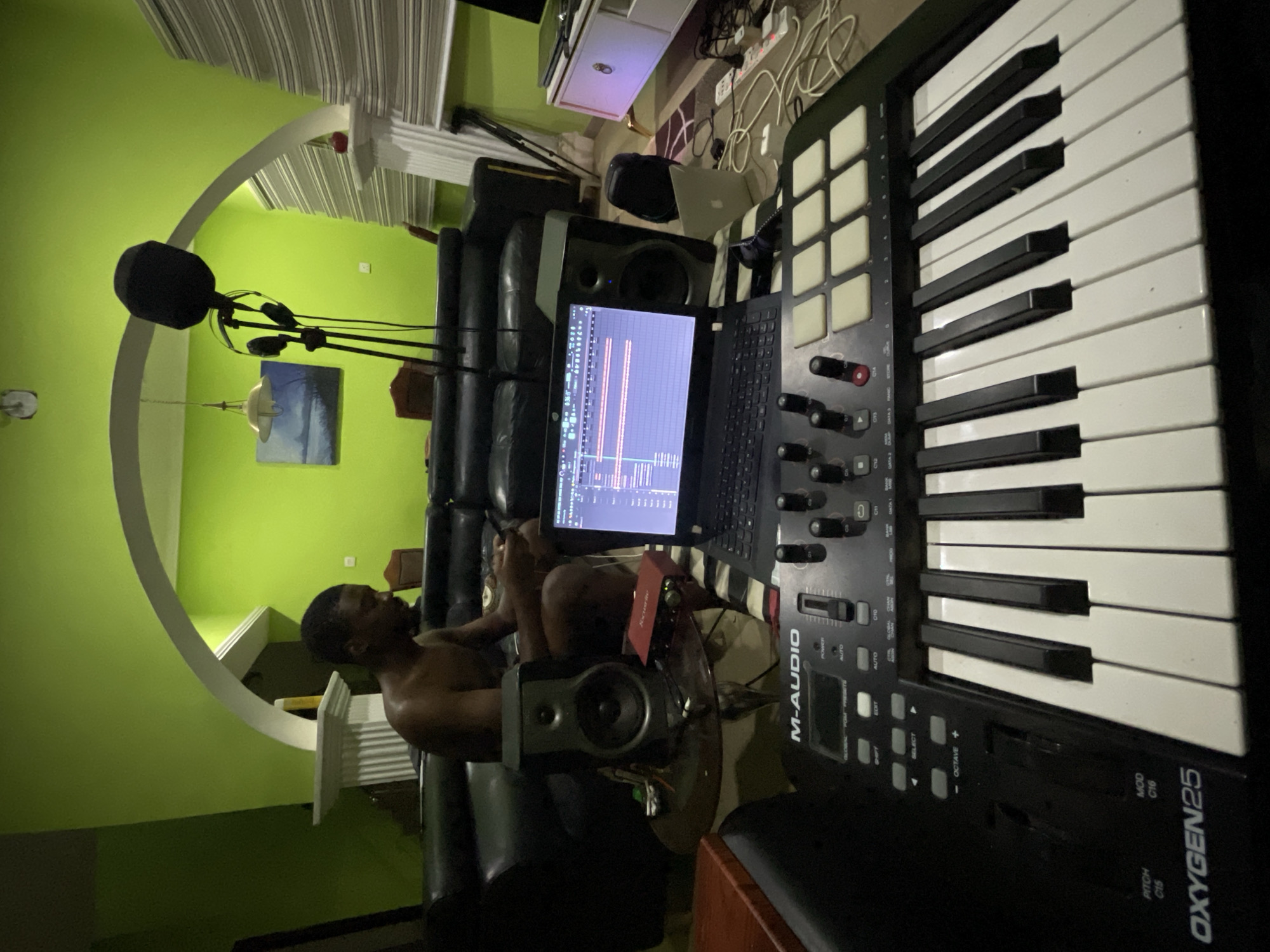The image depicts a man in a recording space. He is sitting in a black leather chair. A laptop with music production software open is in front of him, positioned between studio monitors. In the foreground is an M-Audio Oxygen 25 MIDI keyboard. A microphone with a pop filter is visible in the background. The walls are painted a lime green color. The location may be in Abuja, Nigeria, based on the presence of the man. Other objects visible include a painting, a desk, other sound recording equipment, and a rug.