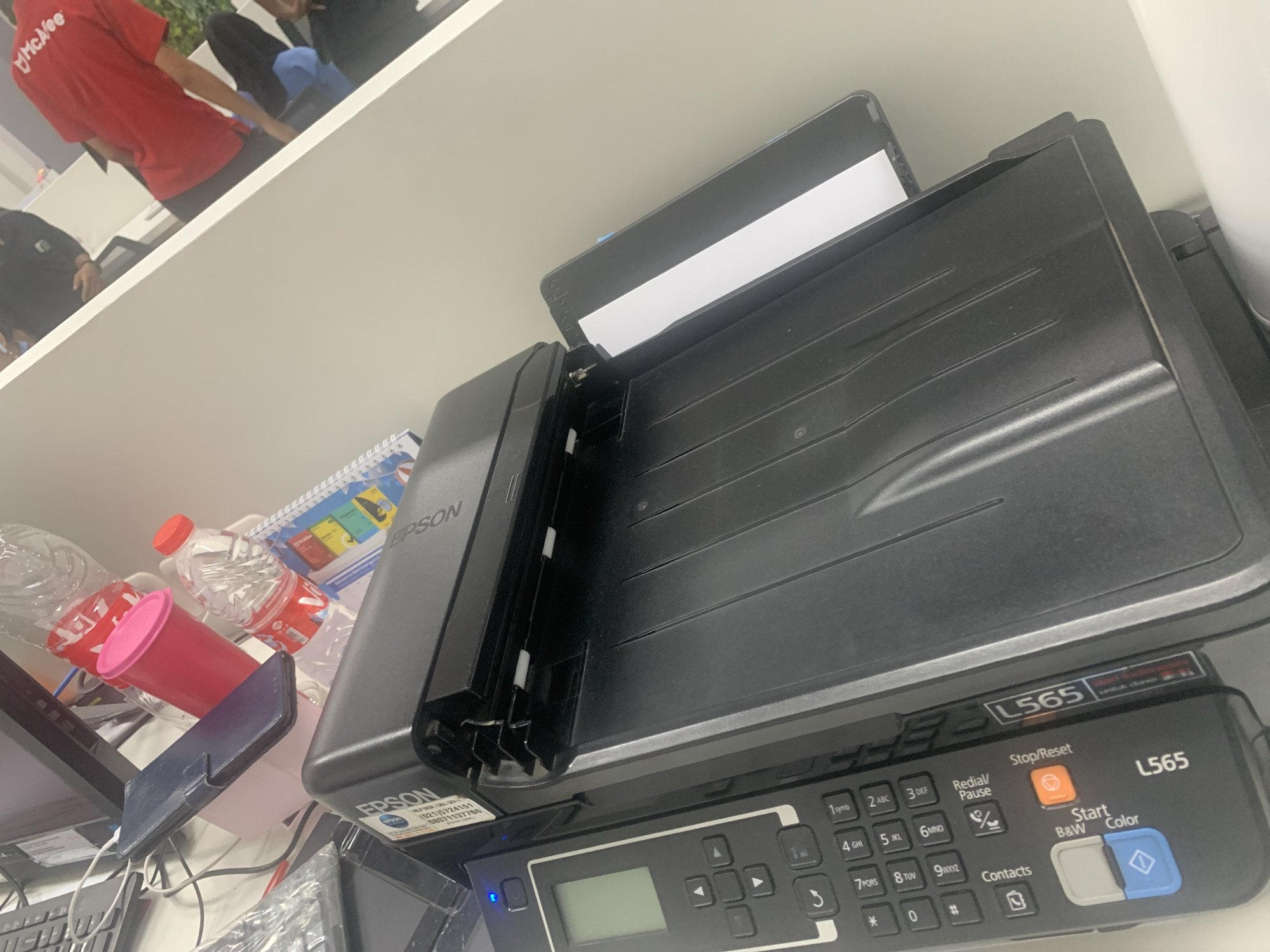A slightly angled, close-up shot shows an EPSON L565 printer on a white desk. The printer is black and has a document feeder on top. There are several buttons and a small LCD screen on the front of the printer. A sheet of white paper is partially inserted into the document feeder. In the background, a red t-shirt with "McAfee" printed on it is visible, worn by a person sitting on a chair. Other objects on the desk include plastic water bottles, a pink cup, and a smartphone. The scene appears to be an office environment.