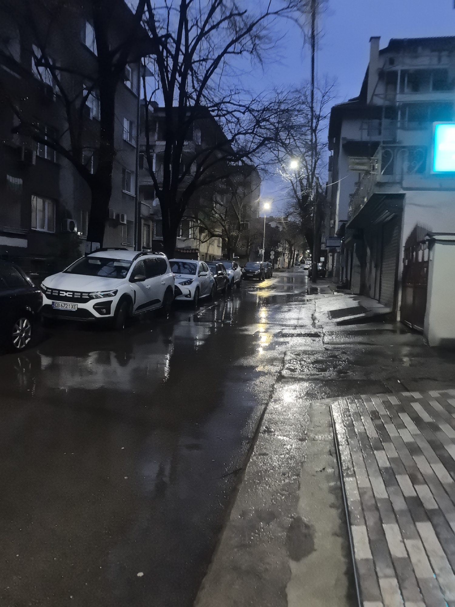 The image depicts a street scene in Stara Zagora, Bulgaria, after rainfall. The street and sidewalks are wet, reflecting the streetlights and the sky's ambient light. Cars are parked along the left side of the street, including a white Dacia with license plate "CB 6721 XE". Buildings line both sides of the street, with bare trees visible between them. The sky is a muted blue, indicating either early morning or late evening. The sidewalks are paved with rectangular gray bricks. The overall impression is a quiet, wet urban environment.