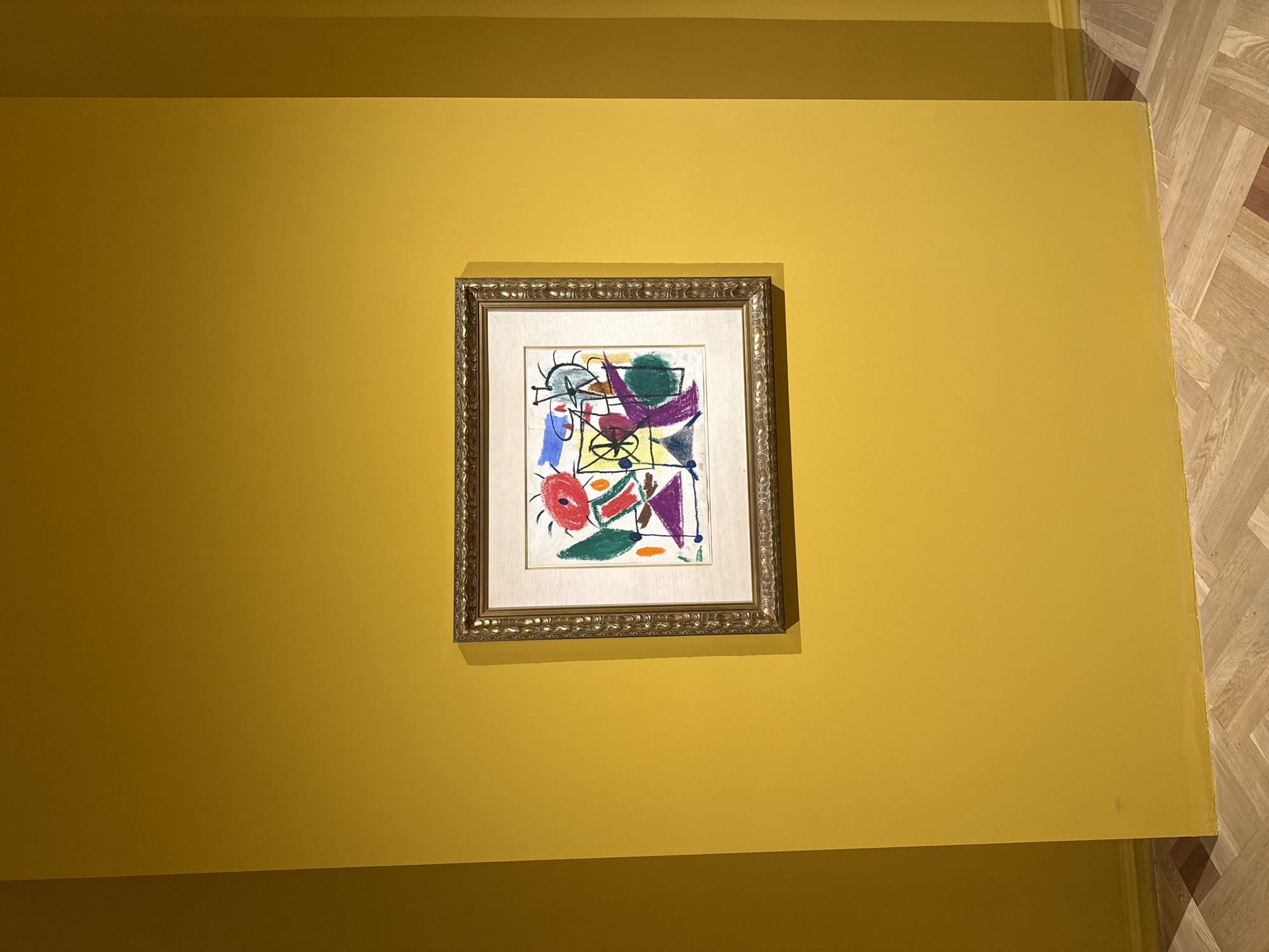 The image depicts a framed abstract painting hanging on a yellow wall. The painting features various geometric shapes and lines in colors such as red, green, blue, purple, and black. The frame is ornate and appears to be gold or a similar metallic color. The wall itself is a solid yellow color. To the right, there's a glimpse of a parquet-style wooden floor, suggesting an interior setting, probably in Yerevan, Armenia.