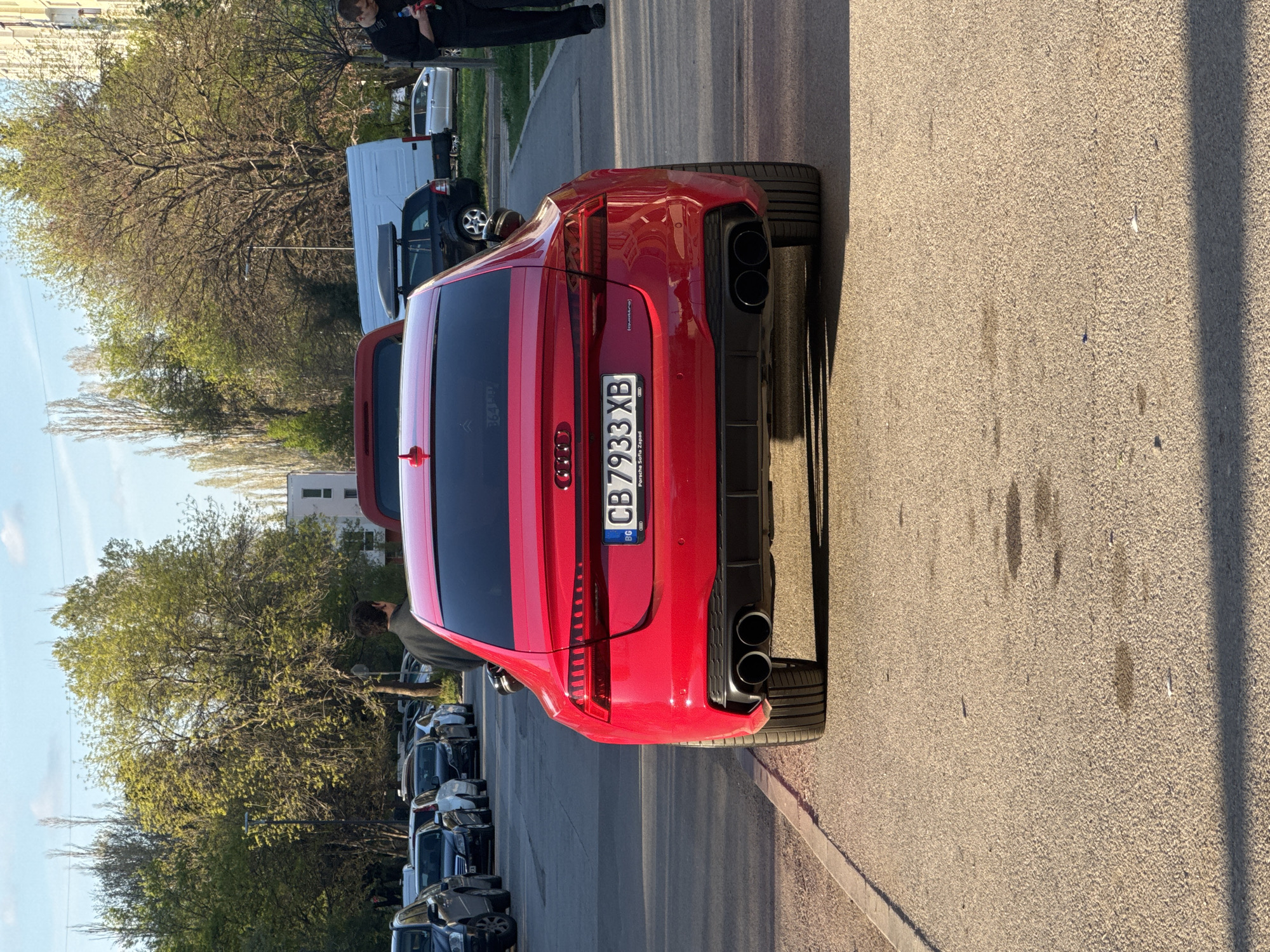 A red Audi RS4 Avant is parked on the side of a street in Sofia, Bulgaria, as indicated by the license plate "CB 7933 XB". The car has a distinct red color, quad exhaust pipes, and a black rear diffuser. A person is standing next to the driver's side. There is a row of parked cars and trees lining the street. A person is visible in the background. The street is paved and the sky is light blue.