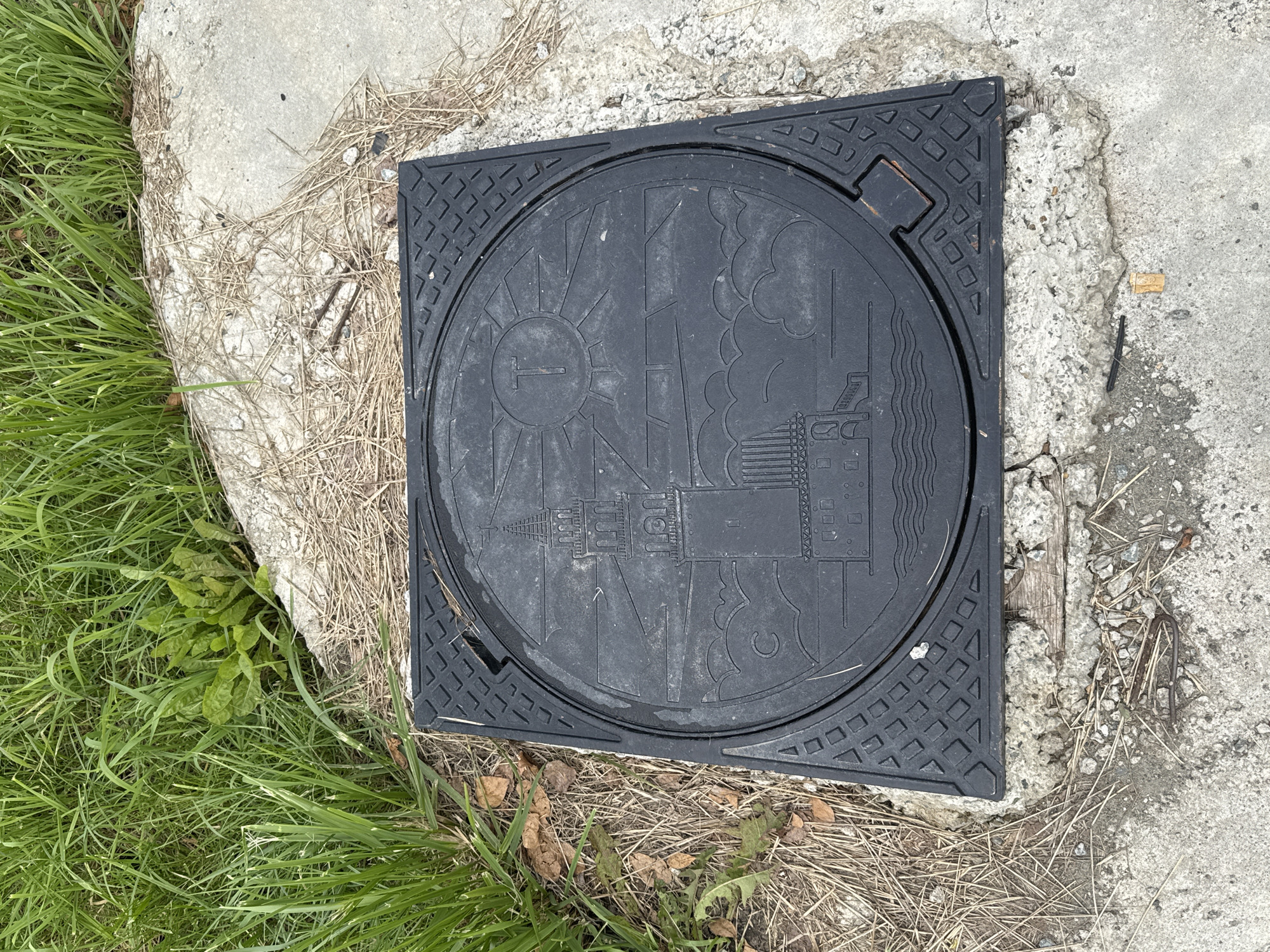 The image shows a square, black metal manhole cover embedded in a concrete surface. The manhole cover has a circular design in relief, featuring a lighthouse with a sun and clouds above it. The lighthouse is depicted with multiple levels and windows. A letter "T" is visible within the sun. The surrounding area of the manhole cover has a textured, geometric pattern. The concrete surface is cracked and weathered, with dry grass and pine needles scattered around it. In the background, there is lush green grass. The manhole cover is positioned in an outdoor environment, likely a public space in the city of Nevyansk, Russia, judging by the artistic depiction of the Nevyansk leaning tower on the manhole cover.