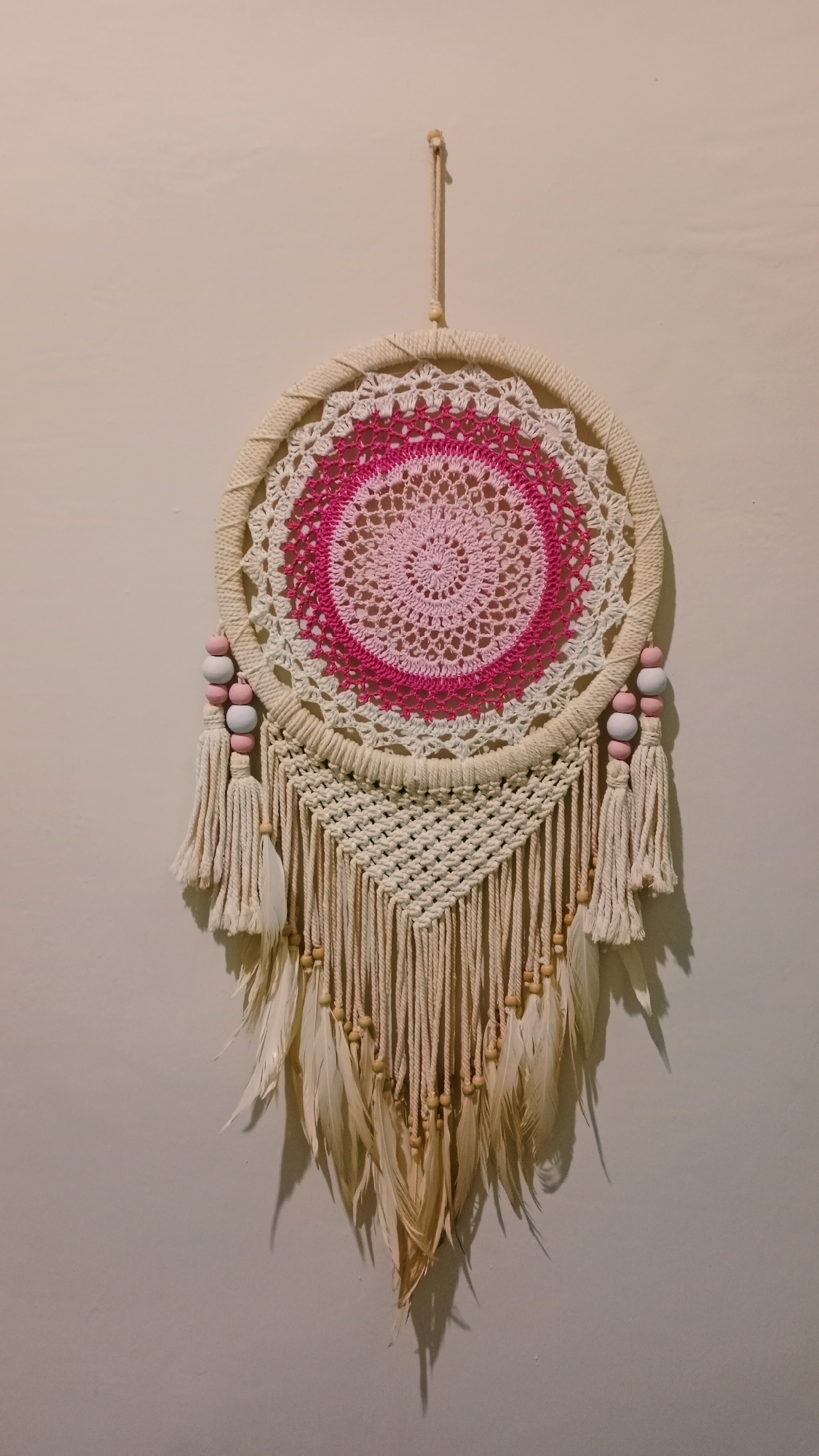 A dreamcatcher hangs against a plain, light-colored wall. The dreamcatcher is circular, with a woven outer ring and a crocheted inner circle featuring intricate patterns in white, light pink, and dark pink. Three cream-colored tassels hang from the bottom of the circular frame, each adorned with light pink and white wooden beads. Below these tassels, a macrame-style fringe with numerous cream-colored cords extends downwards, interspersed with small wooden beads. The bottom of the dreamcatcher is decorated with several cream-colored feathers. The lighting casts a shadow of the dreamcatcher onto the wall. The scene is indoors, with no visible indication of the specific location in Karang Dukuh, Indonesia.