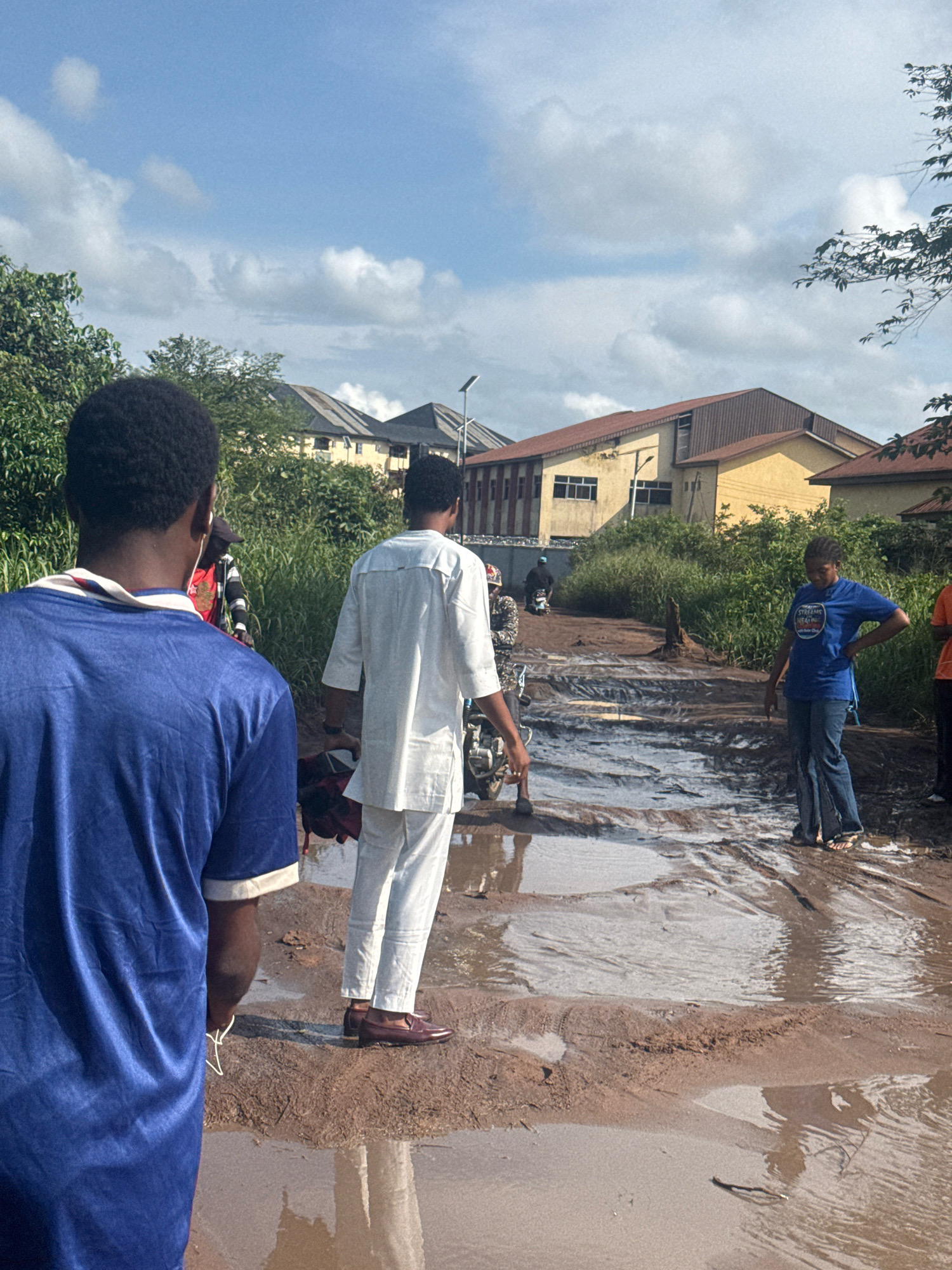 In Ihiagwa, Nigeria, a group of people navigate a muddy, waterlogged road with visible puddles and ruts. In the foreground, a person wearing a blue t-shirt is seen from the back, facing away from the camera. To their right, another individual dressed in white, with a traditional Nigerian outfit, walks towards the camera. Further down the road, a young woman in a blue t-shirt and jeans stands with her hand on her hip, observing the scene. Several motorcycles are present, with one rider visible in the background. The environment is characterized by lush green vegetation on the sides of the road and buildings in the distance. The sky above is a mix of blue with scattered clouds.