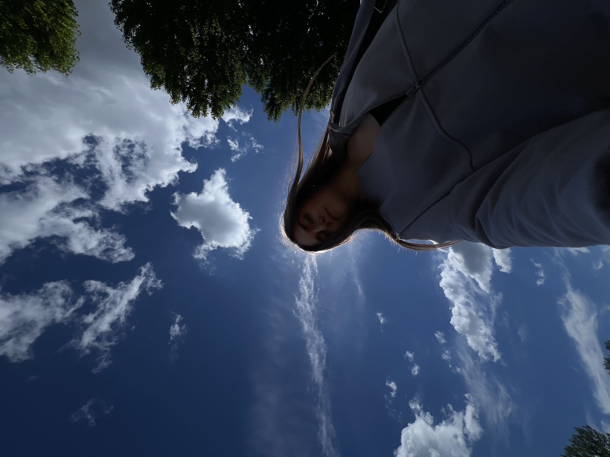 A low-angle, wide-shot, outdoor photograph captures a young woman with long, brown hair standing beneath a vast expanse of blue sky with scattered white clouds. The sun is positioned behind her head, creating a strong backlight effect that illuminates her hair and the edges of her gray jacket. She is looking down and slightly to her right, with her face partially obscured by shadows and the backlighting. Her expression is serene. The woman is wearing a gray zip-up jacket over a dark garment, possibly a tank top or t-shirt. The photograph was taken in Ternopil, Ukraine. The upper right corner of the frame shows the dark green foliage of a mature tree, and the bottom edge of the frame reveals the tops of more trees or greenery. The overall scene conveys a sense of open space and natural light.