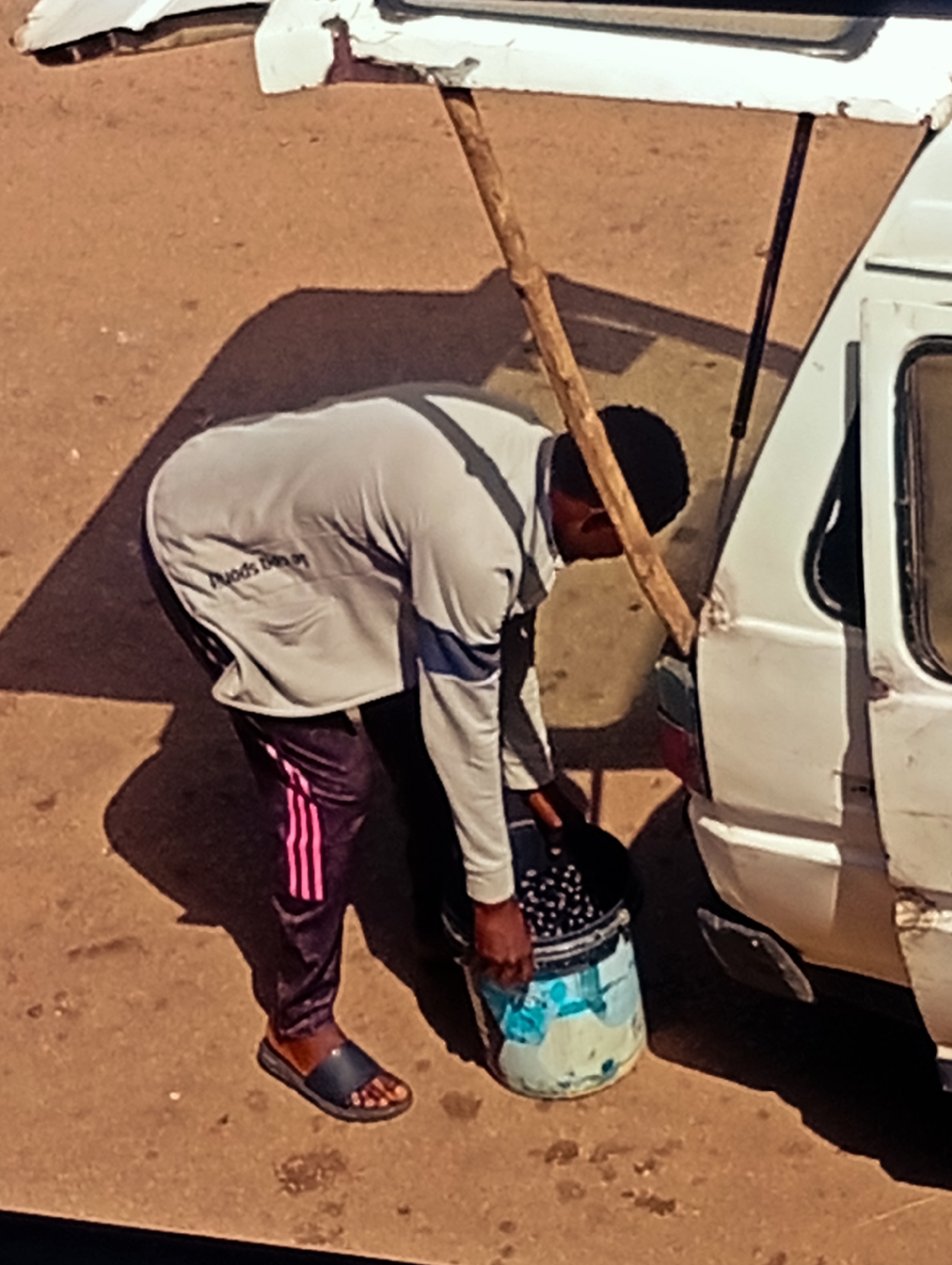 In Bukuru, Nigeria, a person is seen from a high angle view, bent over a large bucket filled with small, dark, round objects, possibly beads or seeds. The bucket is light blue and white with colorful streaks. The person is wearing a grey long-sleeved shirt with a dark blue stripe on the sleeve and dark pants with pink stripes down the legs. They are also wearing dark sandals. A wooden pole leans against a white vehicle parked nearby, its rear quarter panel dented. The ground is a dusty, reddish-brown color, indicative of the soil in the area. A long shadow is cast to the left of the person, suggesting bright sunlight. The scene appears to be an outdoor market or roadside area.