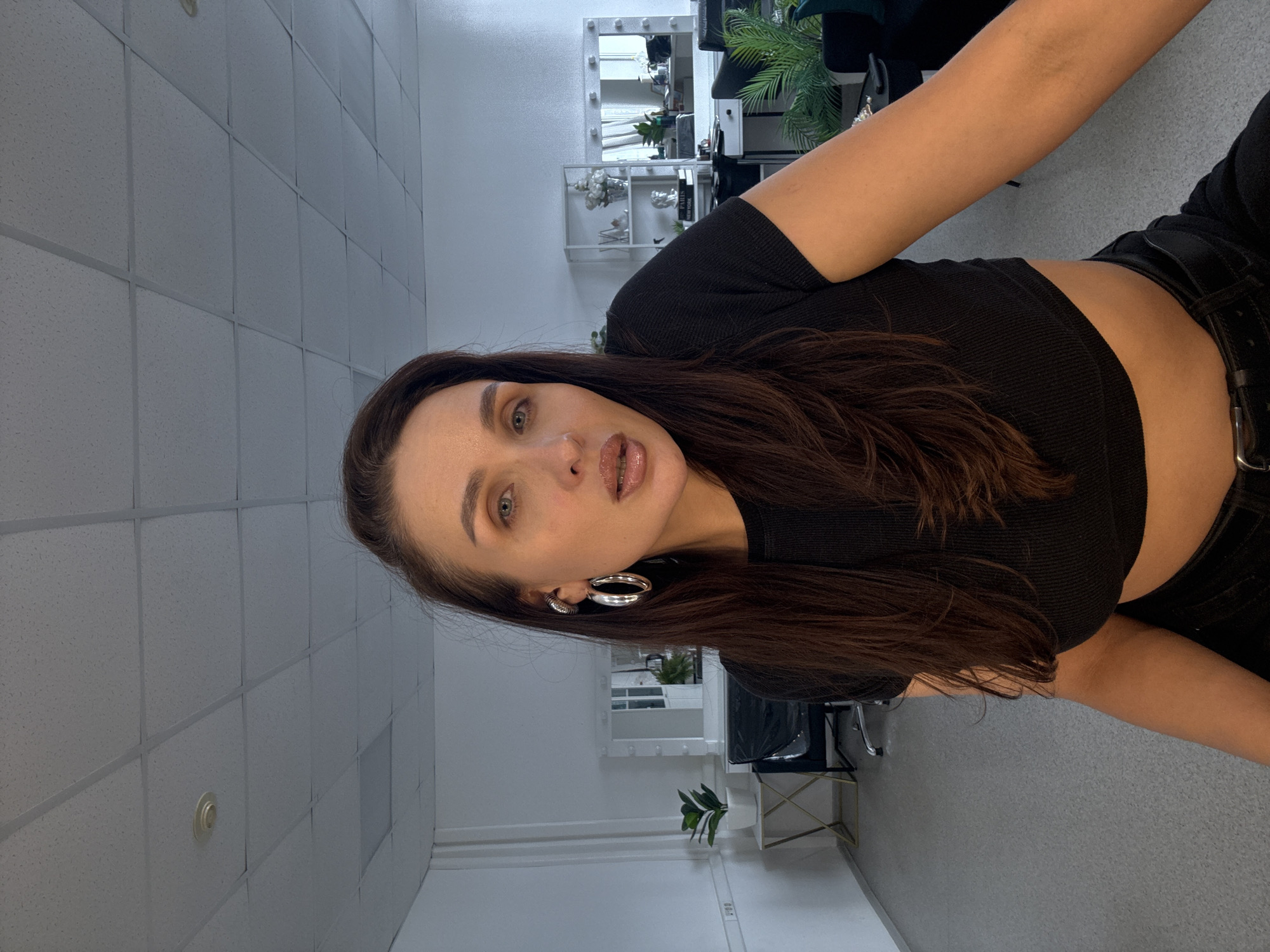 A woman with long brown hair, wearing a black crop top and black pants with a belt, is looking at the camera. She is wearing silver hoop earrings and has light eyes. The background features a room with a drop ceiling, a makeup mirror with lights, plants, and a chair. The location might be a beauty salon or studio. While it is possible the photo was taken in a similar establishment in Chaikovsky, Russia, the photo itself does not contain information to confirm it.