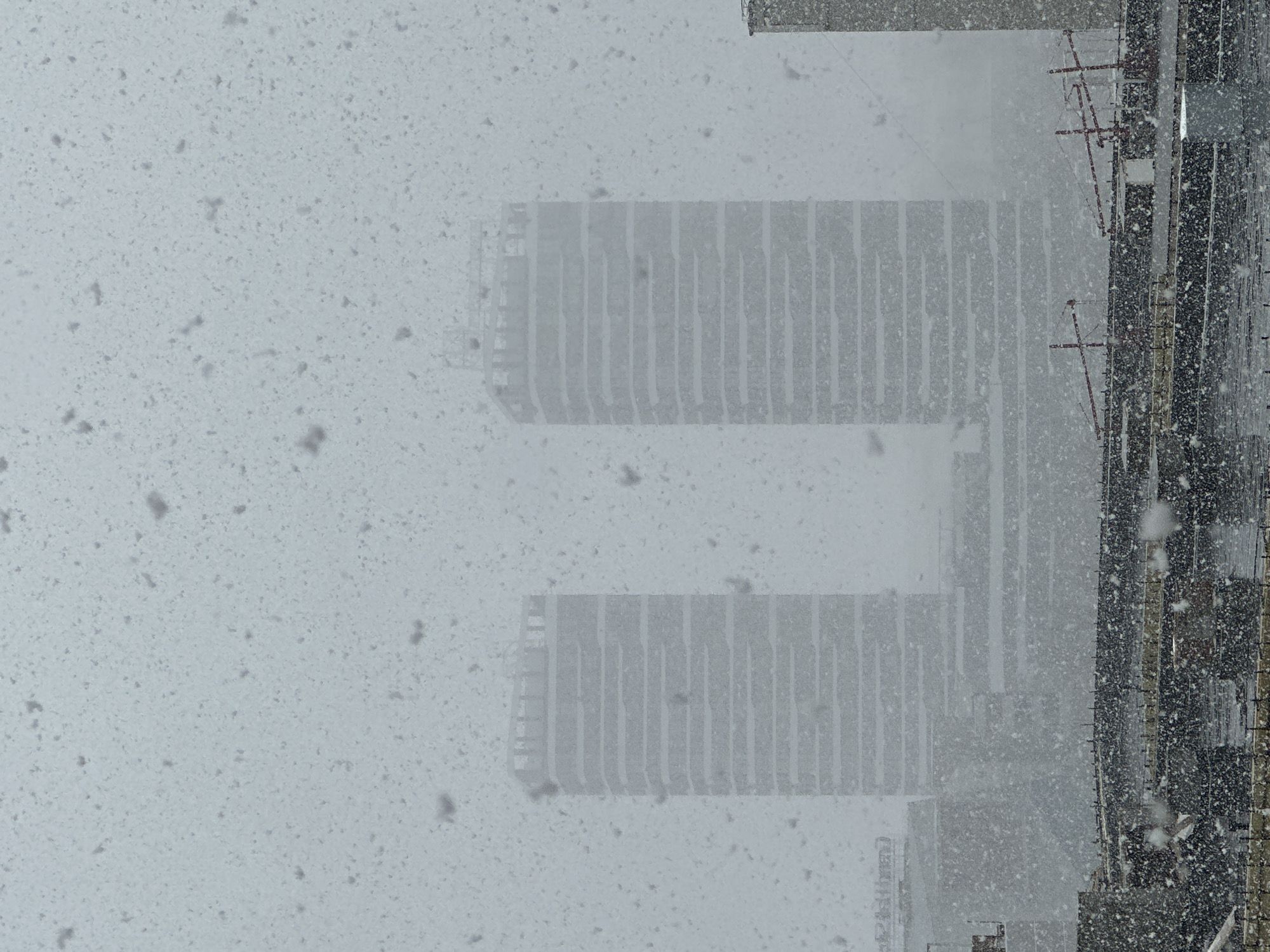 The image, taken during a heavy snowfall, shows an urban landscape with two indistinct multistory buildings in the background. The buildings feature a repetitive linear pattern, possibly reflecting the facade design or balconies. To the right of the buildings, construction or industrial structures are visible. The falling snow obscures the view and covers most of the image. It is strongly indicated that this is of the Afontovo district in the city of Krasnoyarsk, Russia.