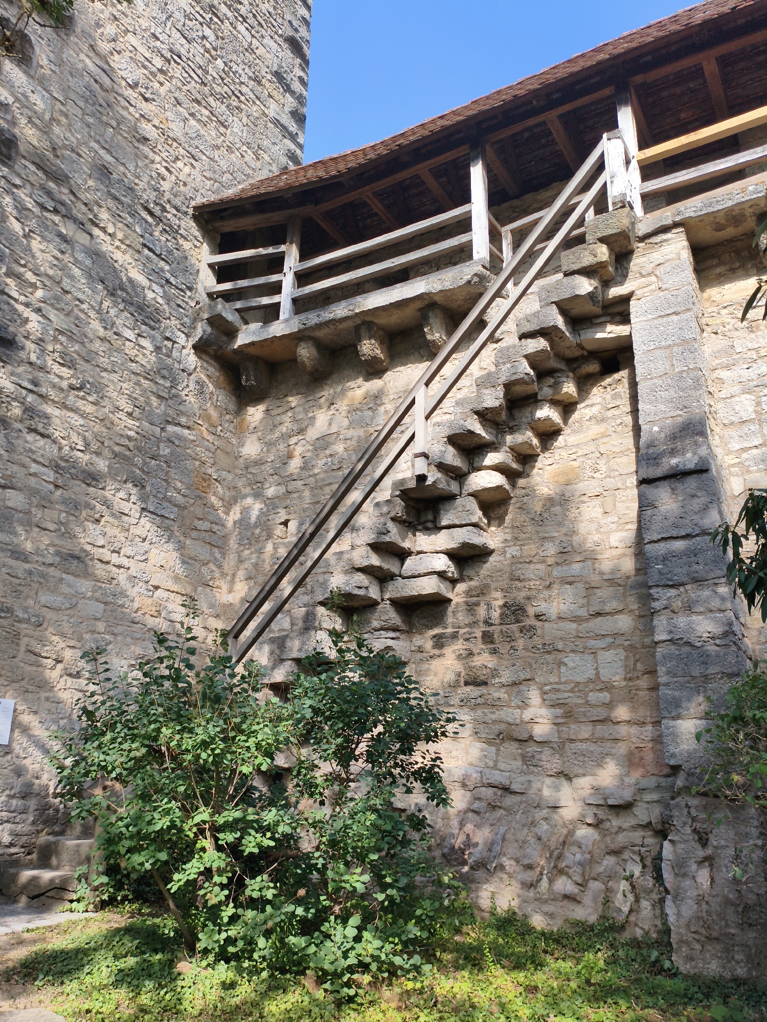 The image depicts a section of the medieval town wall of Rothenburg ob der Tauber, Germany. In the foreground, a lush green bush with small leaves grows on the ground, which is covered with more ground cover plants and dappled with sunlight and shadows. To the left, a set of stone steps is partially visible, leading upwards.
The main focus of the image is the stone wall and an external staircase. The wall is constructed from rough-hewn, irregularly shaped stones of varying sizes and colors, predominantly in shades of beige and gray, with some darker stones providing contrast. Sunlight casts distinct shadows on the textured surface of the wall.
An unusual stone staircase juts out from the wall, featuring individual stone treads that appear to be cantilevered or supported in an unconventional manner. The steps are made of lighter-colored stone and are arranged in a zig-zag pattern. A simple wooden railing with diagonal supports runs alongside the staircase, providing a barrier. Above the staircase and railing, a wooden walkway or covered passage is visible, with a tiled roof composed of reddish-brown tiles. This elevated walkway is supported by wooden beams and pillars.
The sky is a clear, bright blue. The overall impression is one of ancient architecture integrated with natural elements, characteristic of historical European cities.