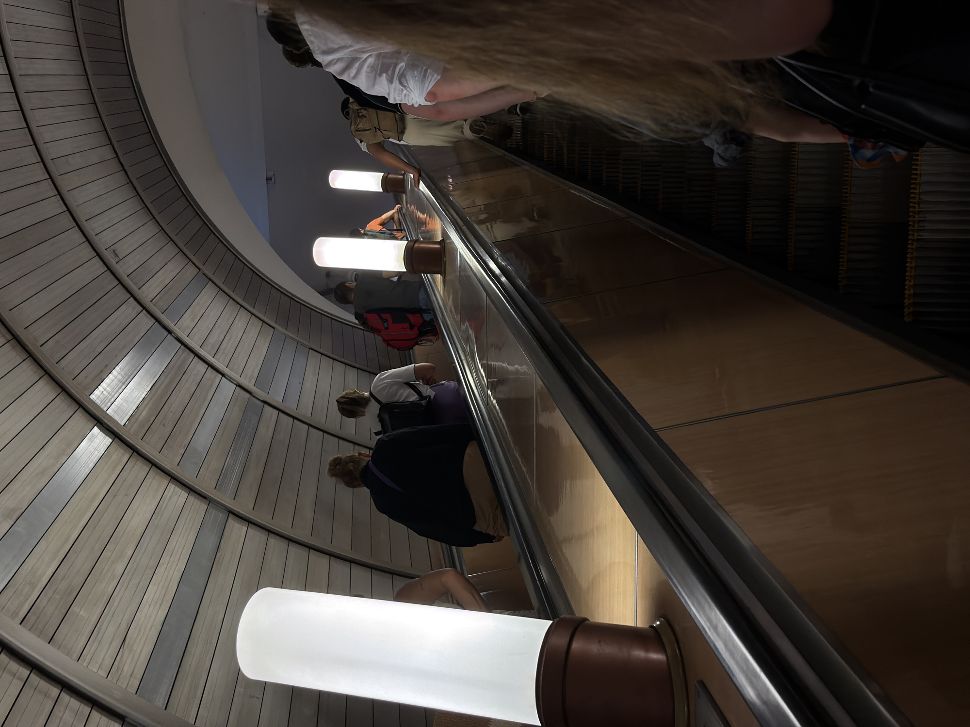 The image captures an upward-moving escalator in a Moscow Metro station, characterized by its distinctive wooden-paneled, arched ceiling. Several individuals are ascending the escalator, some carrying backpacks, suggesting they are commuters or travelers. The escalator steps are dark and metallic, contrasting with the warm, reflective side panels and the bright, cylindrical light fixtures that illuminate the ascent. The overall ambiance is one of urban transit, with a focus on the architectural elements of the station and the flow of people.