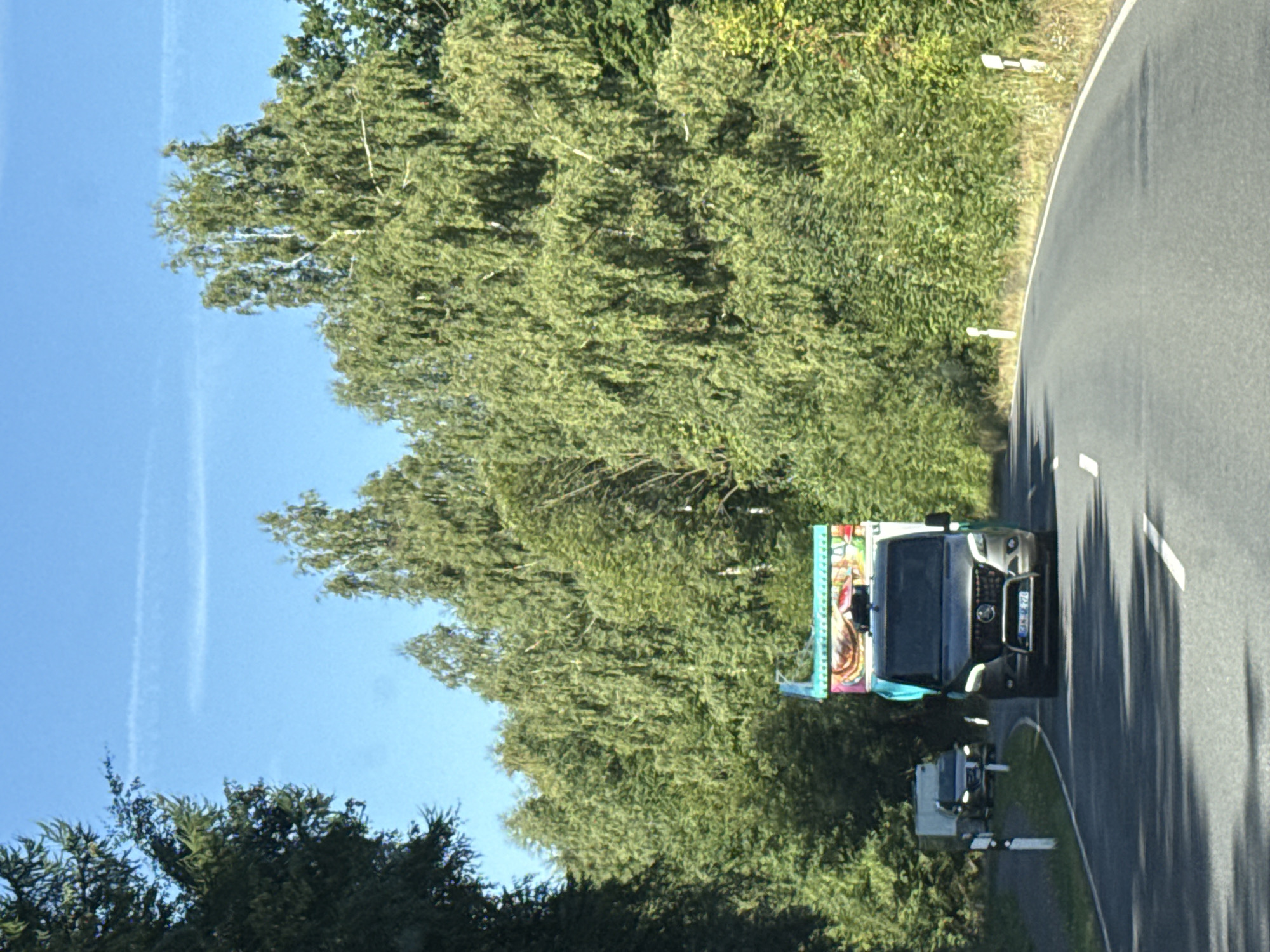 In Walkenried, Germany, on a sunny day, a dark grey van with a colorful, possibly amusement-themed, trailer is driving down a paved road lined with lush green trees. The van has its headlights on and a visible license plate, "GFK JE 774". In the distance, another lighter-colored van is driving in the same direction. The road curves to the right, and road markers are visible on the side. The sky is a clear blue with a faint contrail.