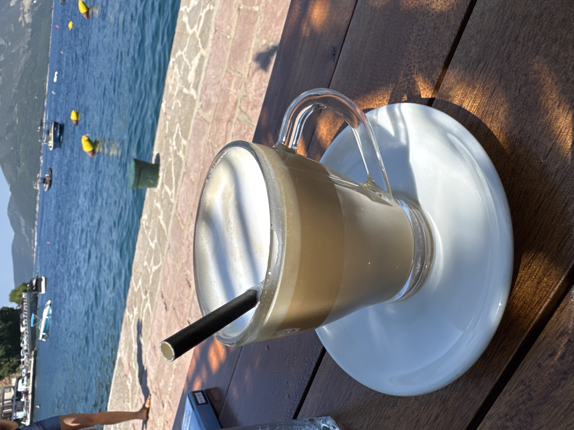 A tall glass of iced coffee with a black straw rests on a white saucer on a wooden table. The coffee has distinct layers of brown and white foam, suggesting it's a latte or macchiato. Sunlight casts dappled shadows on the table and the glass, indicating it's daytime.
In the background, a body of water with small boats and yellow buoys is visible. The water is a vibrant blue, and the shoreline is paved with light-colored stones. Beyond the water, mountains can be seen under a clear, bright sky, suggesting a warm, sunny day. The overall scene depicts a leisurely moment, possibly at a cafe or restaurant by the water. There is no discernible text in the image.