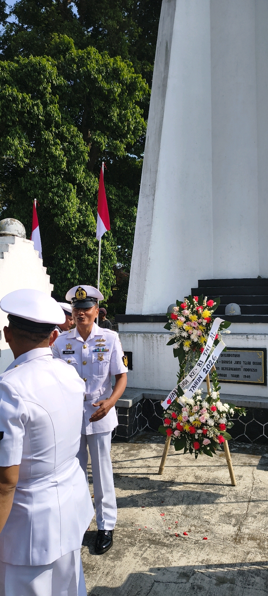The image depicts an outdoor scene in front of a white monument. Two men in white naval uniforms are the primary subjects. The man on the right, identified by the name "BASJDI" on his uniform, is speaking and gesturing with his right hand. He wears a white cap with a naval insignia, a white jacket with medals on the left chest, and white trousers. The other man, on the left, is facing away from the camera, also in a white naval uniform with a white cap. In the background, two Indonesian flags are visible, one to the left and one to the right of a tall, white, triangular monument. A floral wreath with ribbons and flowers in red, yellow, and pink is placed in front of the monument. The ground is paved with concrete, and there are scattered red rose petals. The environment appears to be a commemorative site. The inscription on the monument mentions "HARI JADI KE - 81 TNI AL" and "TAHUN 2026", indicating a celebration for the 81st anniversary of the Indonesian Navy in 2026. The overall atmosphere is formal and commemorative. The location is specified as the city of Sadamantra, Indonesia.