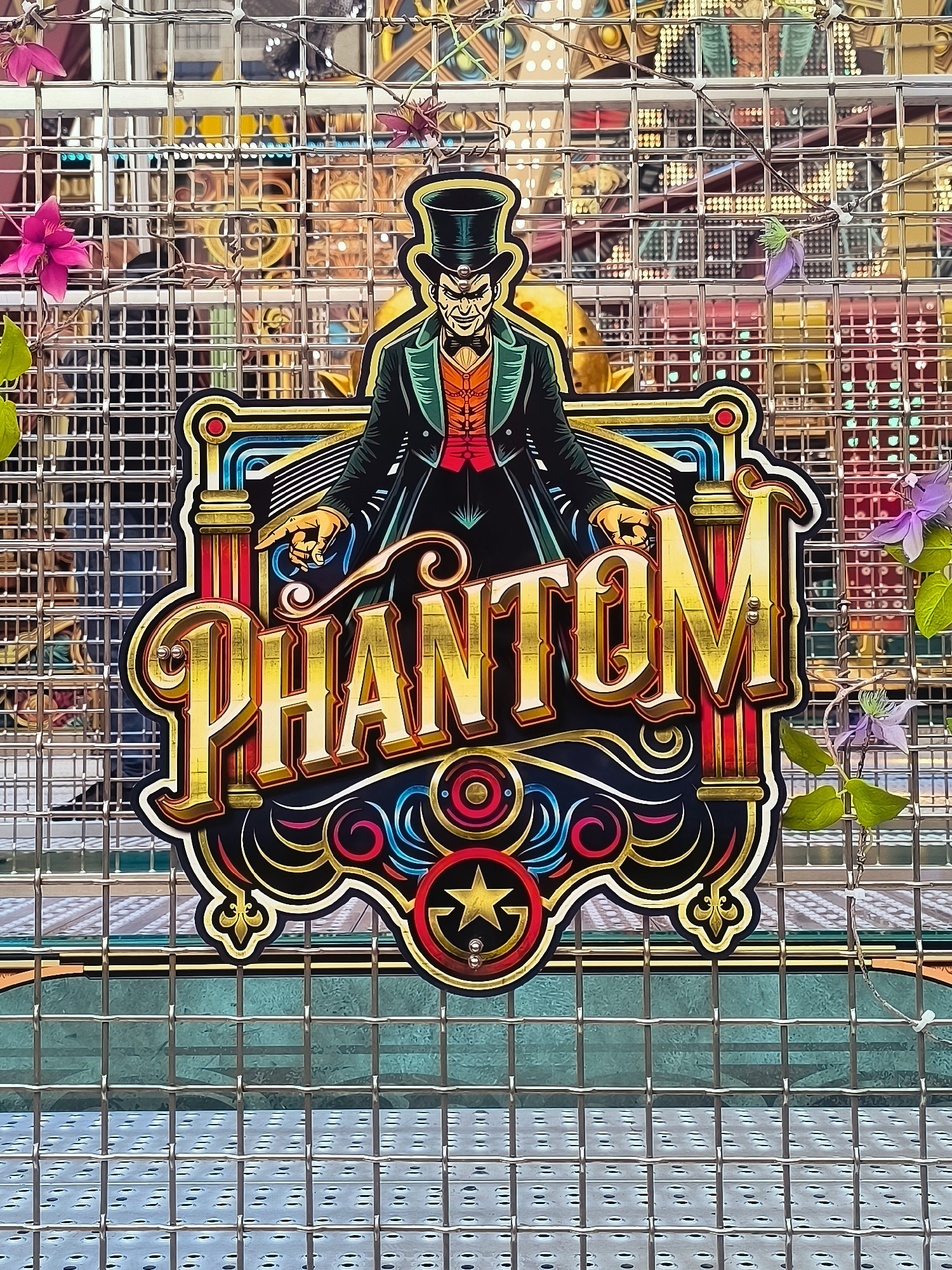 The image shows a close-up of a colorful, stylized sign for "PHANTOM" which appears to be part of a carnival attraction or game. The sign features a cartoonish depiction of a sinister-looking man in a top hat and formal attire, pointing outwards. The lettering of "PHANTOM" is bold and decorative, with a metallic gold finish. The sign is mounted on a metal grate, which is the predominant background element, and some faint greenery and small purple flowers are visible, suggesting an outdoor setting. Behind the grate, there are blurred lights and structures that suggest a carnival or amusement park environment. The sign is positioned centrally within the frame. The image does not contain any identifiable landmarks or features that would indicate its specific location within Hoorn, Netherlands.