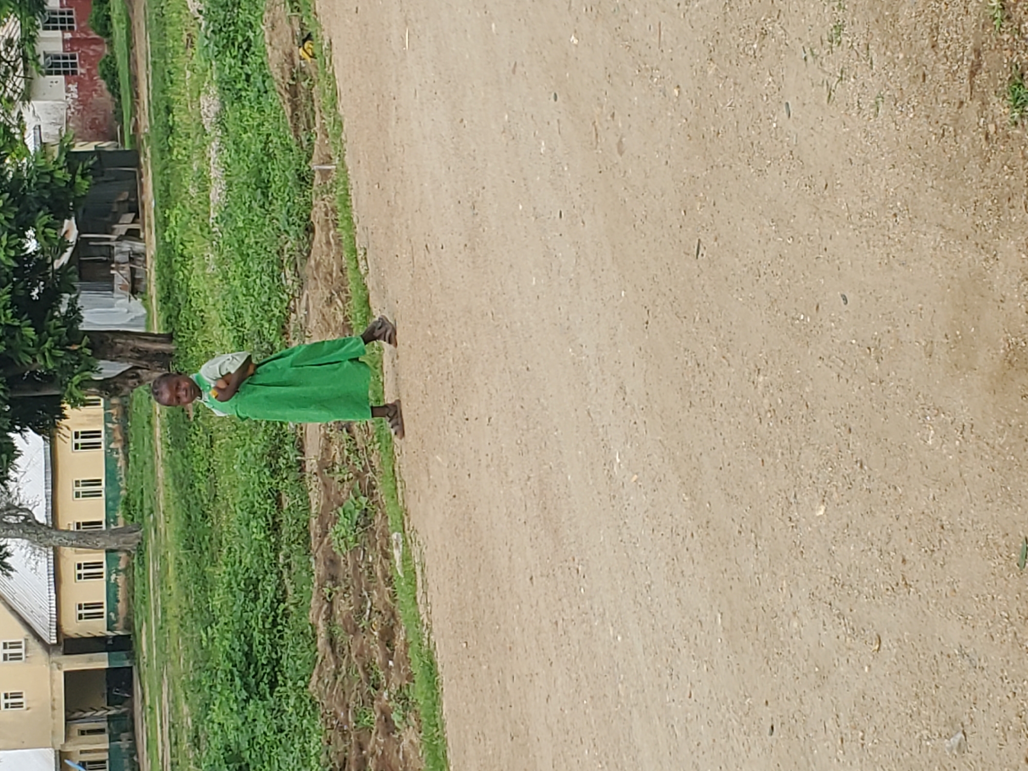 A young child wearing a green dress and dark sandals stands on a dirt road in front of a grassy area and buildings. The child is facing the camera, with their arms held slightly away from their body and their head tilted down. The child's expression appears to be one of sadness or apprehension. The background shows a low-rise building with a light-colored facade and a dark-colored roof. There are trees and vegetation visible in the background as well. The road is dusty and appears to be unpaved. The lighting suggests it is daytime.