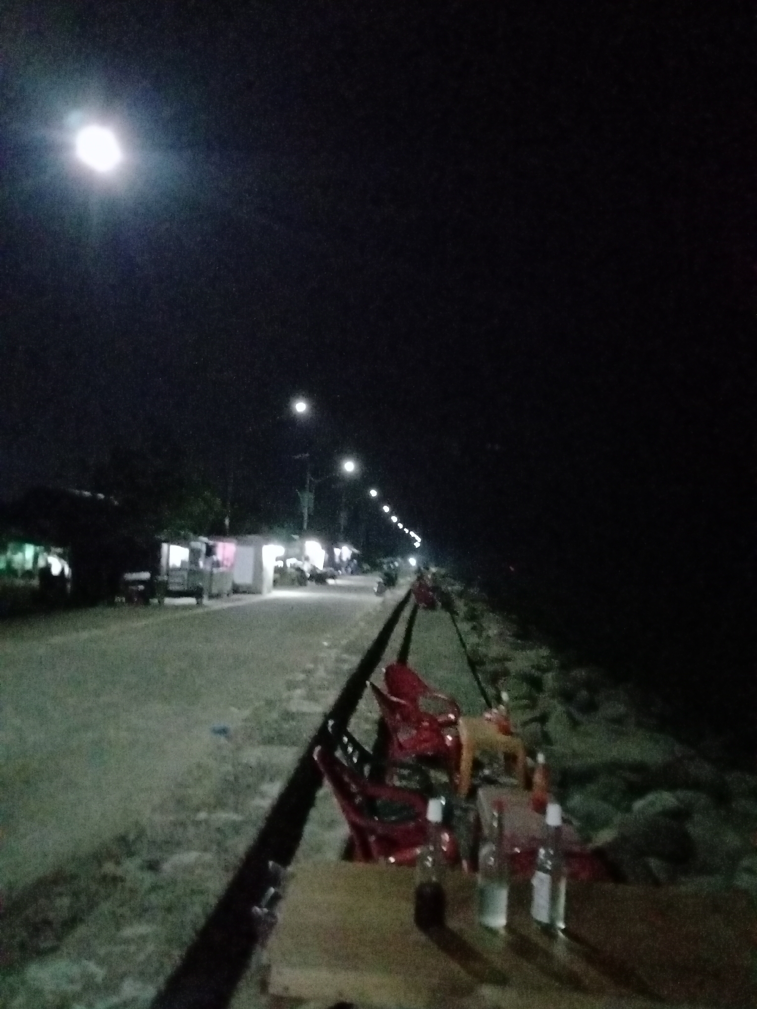 The image depicts a dimly lit street scene at night, potentially in Lhokseumawe, Indonesia. A row of streetlights illuminates a road lined with small buildings or stalls on the left side of the frame. On the right side, there appears to be a shoreline or a breakwater of rocks. In the foreground, a table with bottles on it and some red plastic chairs are present. The general atmosphere suggests an outdoor gathering or public space at night.