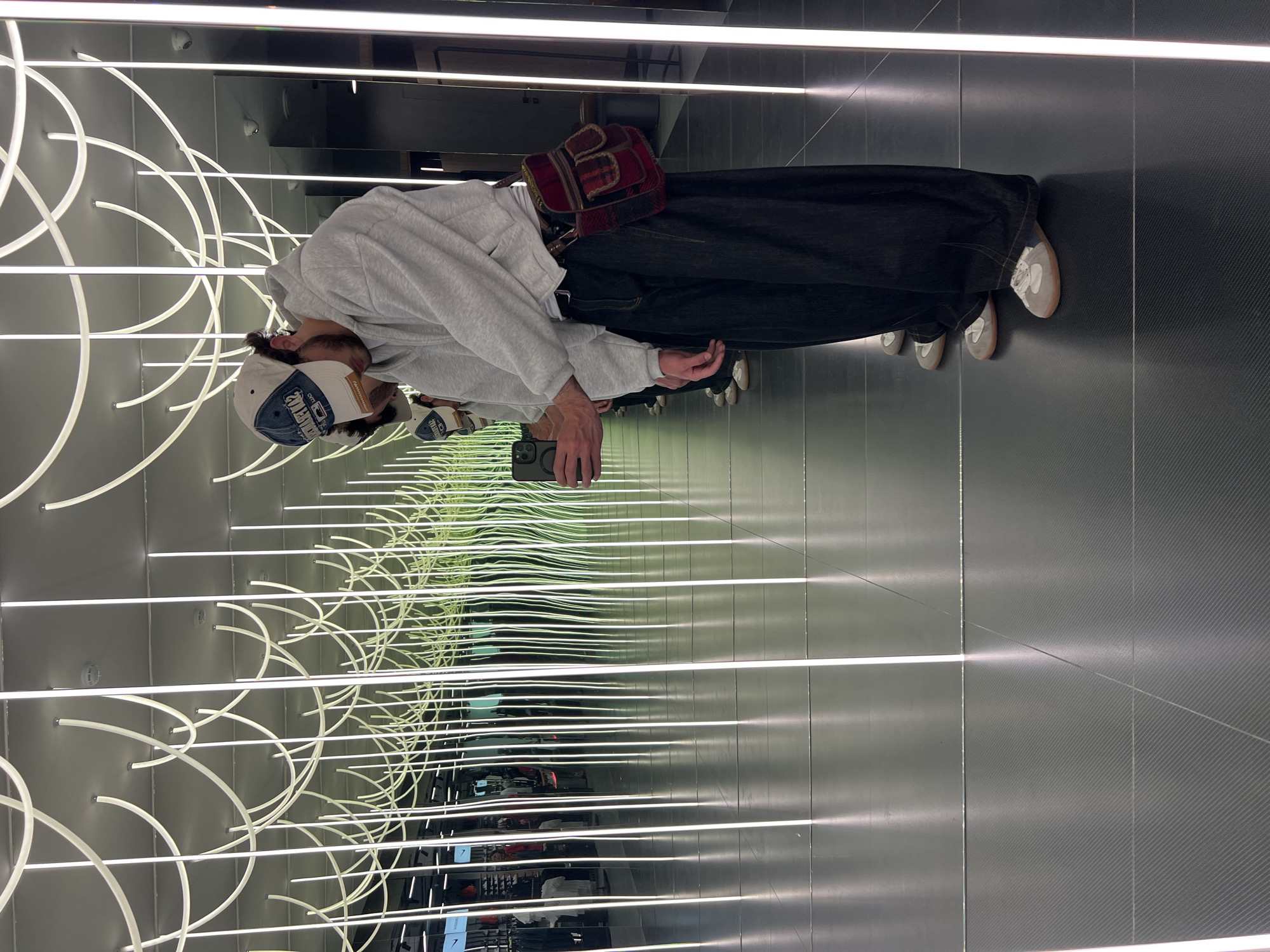 A person is standing in a narrow hallway with mirrors on both sides, creating an infinity mirror effect. The person is wearing a gray hoodie, dark jeans, and gray sneakers. They have a red and brown plaid bag slung over their shoulder. They are holding a smartphone in their hands and looking down at it. The ceiling is adorned with multiple curved white neon lights. The hallway floor and walls are dark gray with a textured pattern. There is no definitive information about the location being Yerevan, Armenia.