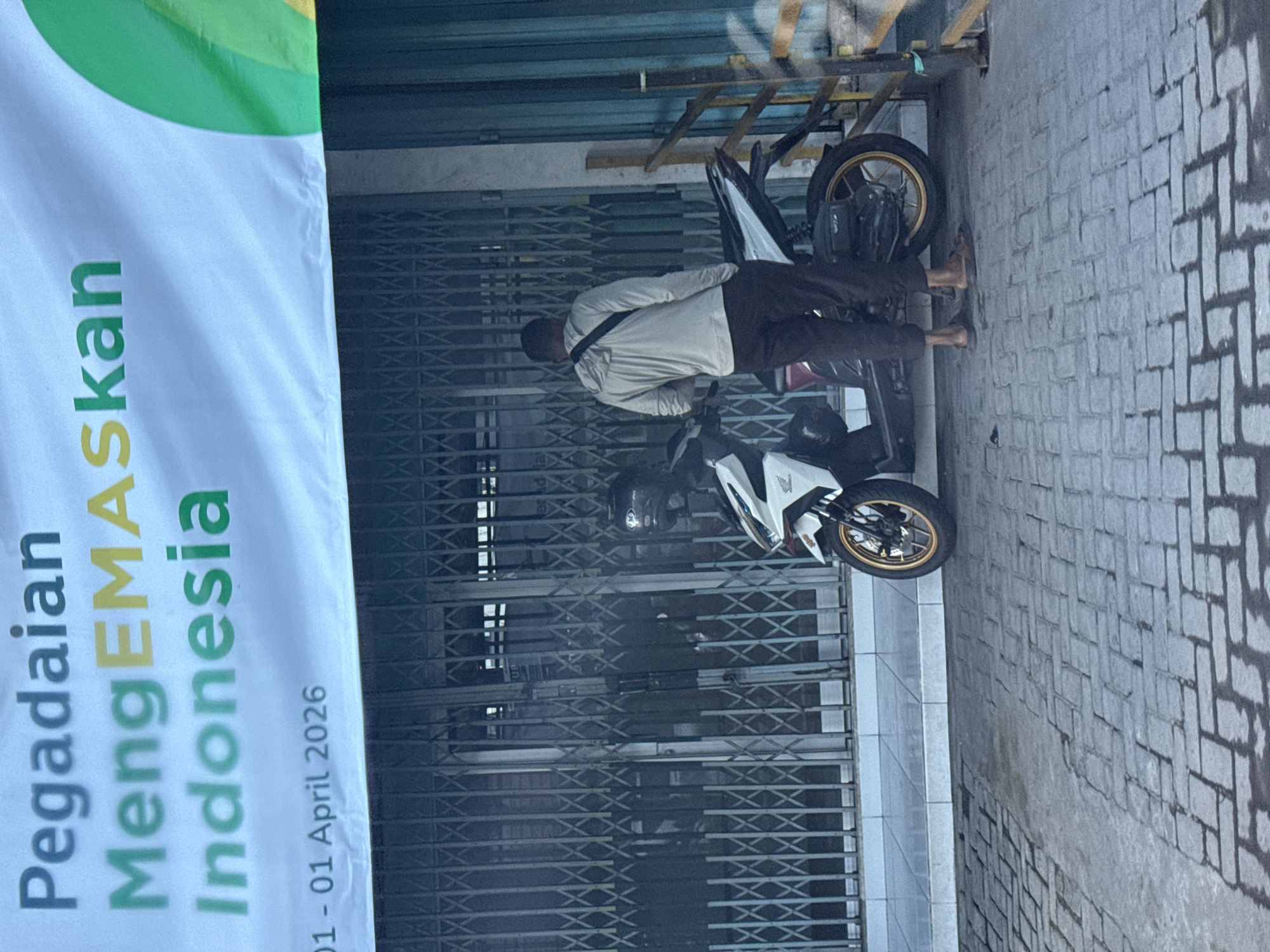 The image, taken in North Kuta, Indonesia, shows an outdoor street scene with a man, two motorcycles, and a promotional banner. A man, wearing a light-colored long-sleeved shirt, dark trousers, and carrying a shoulder bag, stands barefoot near a white Honda scooter with gold-colored wheel rims. Another motorcycle is partially visible behind the man.
The background features the closed metal roller shutter door of a building, suggesting a commercial establishment. To the left, a large white banner is prominently displayed. Its text, in green and yellow, reads: "Pegadaian MengEMASkan Indonesia" followed by "01 - 01 April 2026." This indicates a promotional period or event by Pegadaian, an Indonesian state-owned company.
The ground is a paved sidewalk, characterized by an interlocking brick pattern. The lighting suggests it is daytime, possibly under an overcast sky, as there are no harsh shadows. The overall setting depicts a typical urban or commercial street environment in Indonesia.