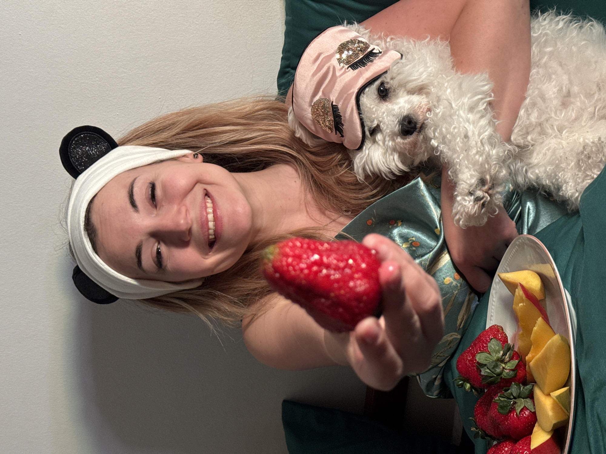 A woman with long, light brown hair and a white headband with black ears is smiling and holding a strawberry toward the camera. She is wearing a green silk pajama top. In her arms, she holds a white, curly-haired dog with a pink eye mask. A bowl of strawberries and sliced mangoes rests on her lap. The setting appears to be indoors with a light gray wall in the background. There is no indication that the image was taken in Budva, Montenegro.