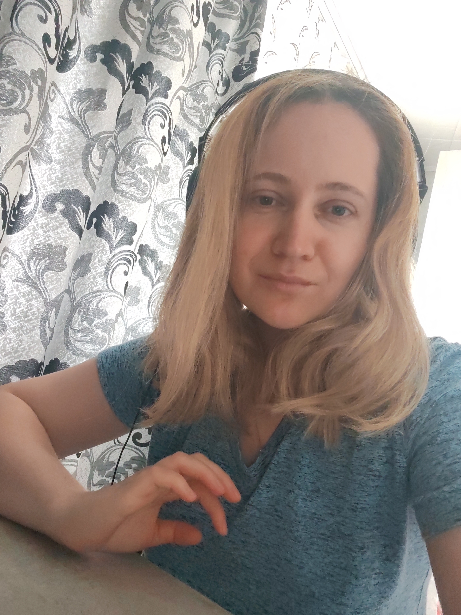 A woman with shoulder-length blonde hair, wearing headphones and a teal v-neck t-shirt, is captured in a close-up, front-facing selfie. She is seated at a table, with her left arm resting on the table and her left hand gesturing slightly. Behind her, a patterned curtain with grey and black floral designs is visible. The lighting suggests it is daytime, likely indoors. The location of this image is unknown, and there is no information to suggest it is located in the city of Сокол, Russia.