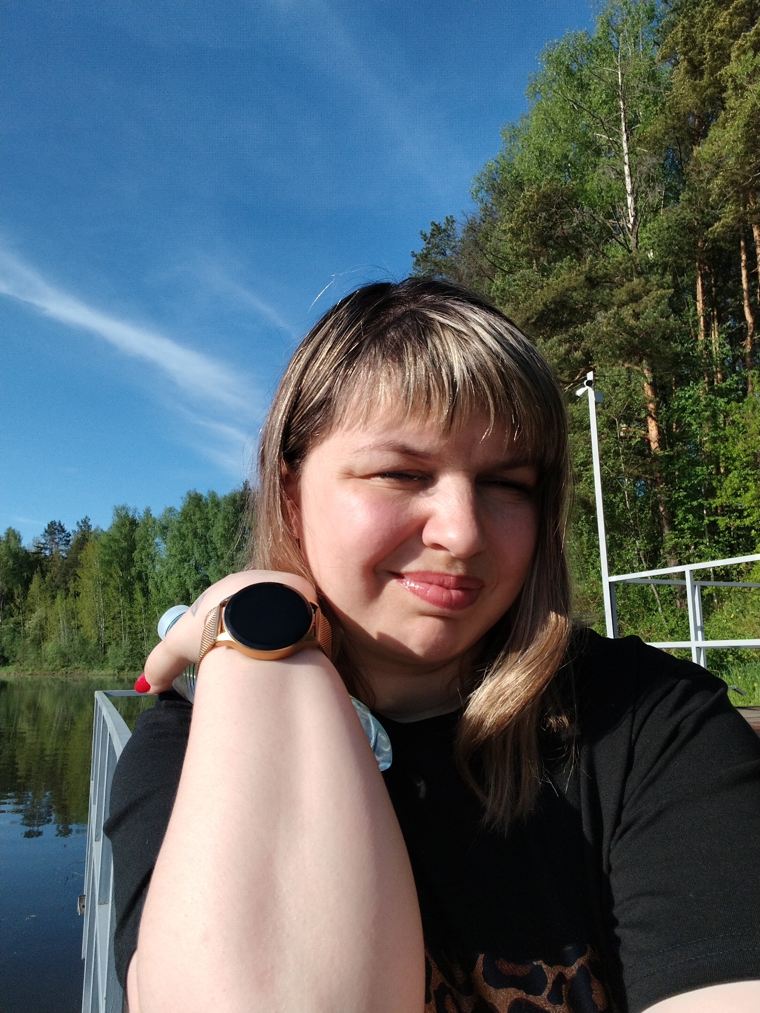 A medium-shot selfie of a fair-skinned woman with light brown and blonde hair, wearing a black top with an animal print design visible, and a gold-colored smartwatch on her left wrist, is taken outdoors. The background consists of a clear blue sky with faint white clouds, a body of water, and a forest. A white railing is also visible in the background. The woman is smiling slightly. The image was taken in the city of Stroitel', Russia.