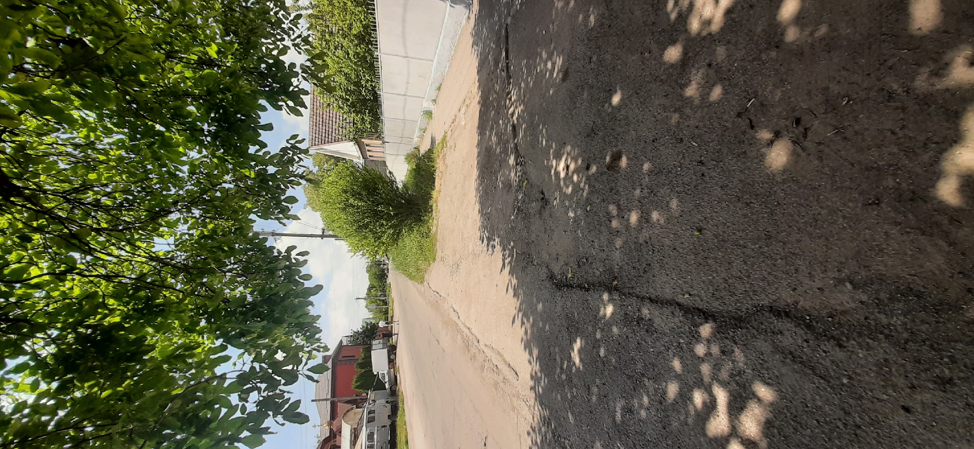 The image depicts a street scene in Yessentukskaya, Russia, on a sunny day. The foreground is dominated by an asphalt road with visible cracks and patches, dappled with the shadow of tree leaves overhead. To the left of the road, several vehicles are parked, including a white van and a smaller white truck, with a red-roofed building in the background. Further down the road, two individuals are walking. On the right side of the road, a light-colored concrete fence runs along a property, with a dense green bush in front of it and a house with a light-colored roof visible beyond the fence. The sky is partly cloudy, indicating fair weather.