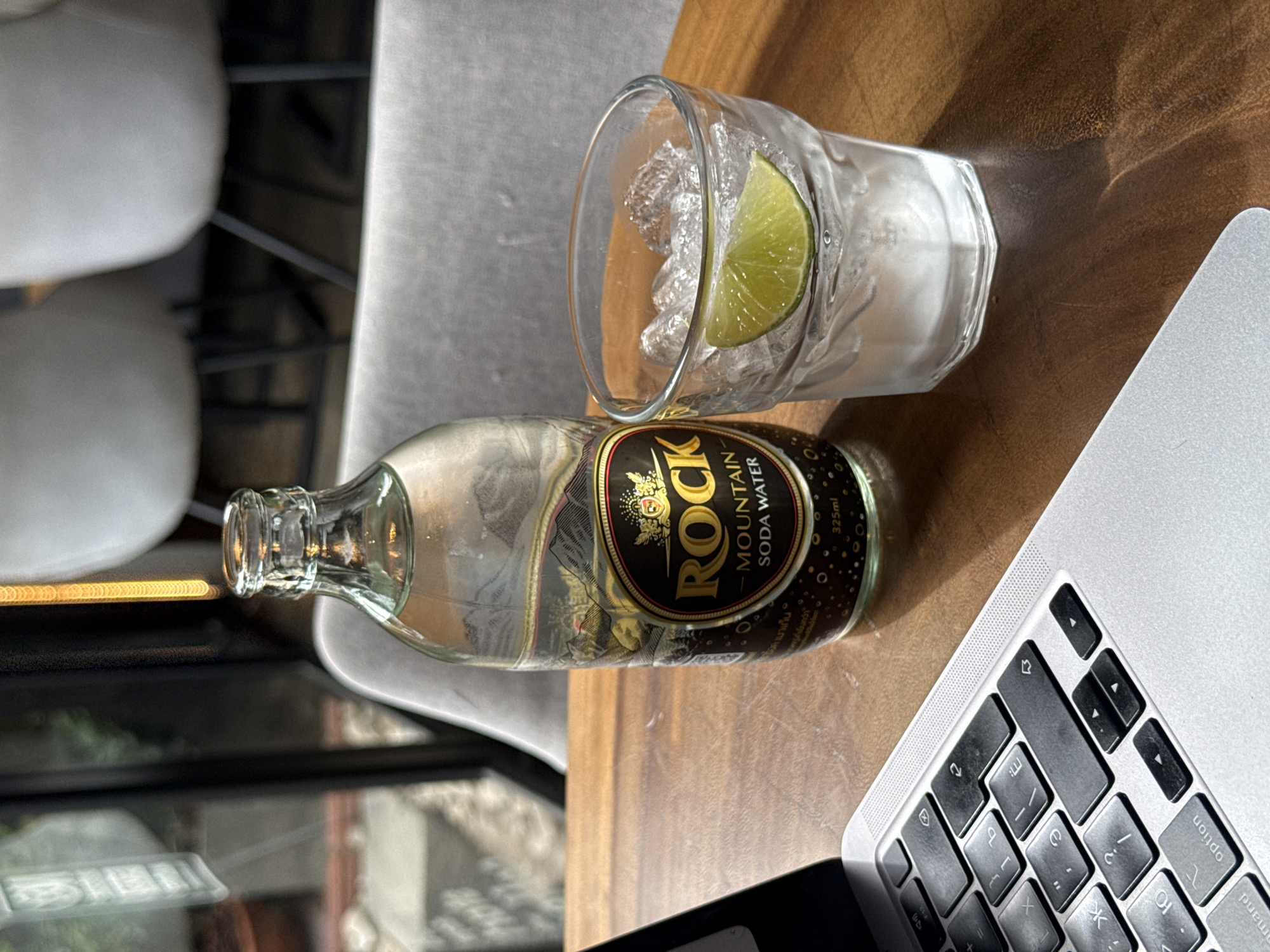 A bottle of Rock Mountain Soda Water and a glass with ice and a lime wedge sit on a wooden table next to an open laptop. The bottle is a clear glass bottle with a black and gold label featuring a mountain landscape. The glass is clear and slightly frosted from the ice. The laptop is silver with black keys. The background shows blurred chairs and a window with a view of greenery. The scene appears to be in a cafe or restaurant setting in Baan Bo Phut, Thailand.