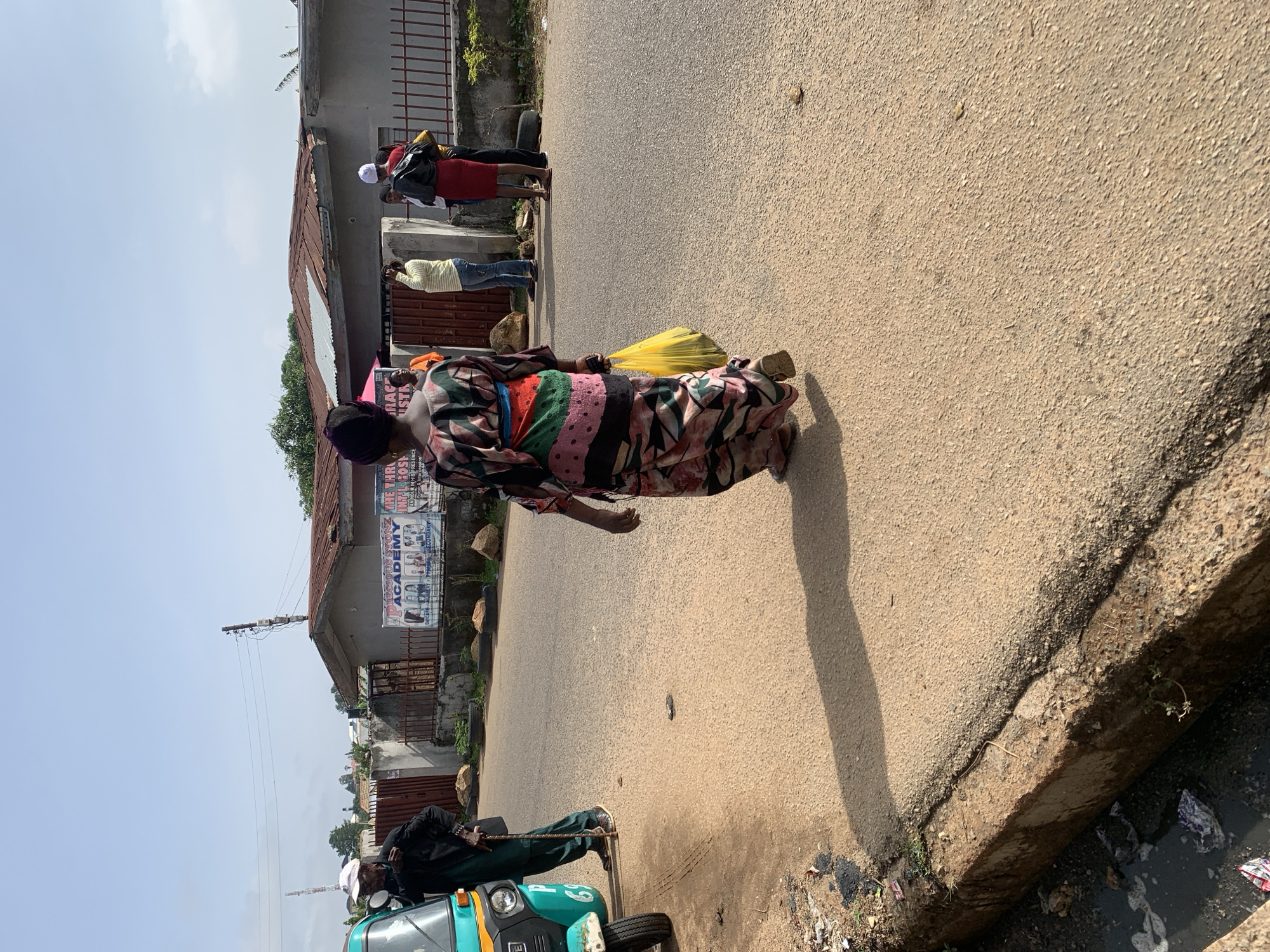 The image captures a vibrant street scene in Jos, Nigeria, during bright daylight. The sky is clear, and strong shadows indicate abundant sunlight. The perspective is from the roadside, looking down a paved road bordered by buildings on the left and a ditch/drain on the right.
Several individuals are visible, engaged in daily activities. In the foreground, a person wearing a multi-patterned dress and a headscarf walks away from the camera, carrying a yellow plastic bag. Further down the road, another person in a yellow striped shirt and blue jeans stands, while behind them, a figure in a red dress with a white head covering is also present. In the bottom left, a person dressed in dark clothing, possibly green trousers and a black top, stands beside a teal and black auto-rickshaw, commonly known as a Keke NAPEP. This individual appears to be holding a cane or stick.
The buildings lining the street exhibit corrugated metal roofs and concrete walls, with some featuring metal gates. A utility pole with overhead wires is also visible. The roadside next to the ditch contains some rocks and debris.
Visible text on a banner attached to one of the buildings reads: "DAYCAR" (likely "DAYCARE" or similar), "ACADEMY INTEL GOSK," "AN HOUR IN HIS PRESENCE," and "SECONDARY." These fragments suggest a school or religious institution. The presence of the auto-rickshaw and the architectural style are consistent with urban environments in Nigeria.