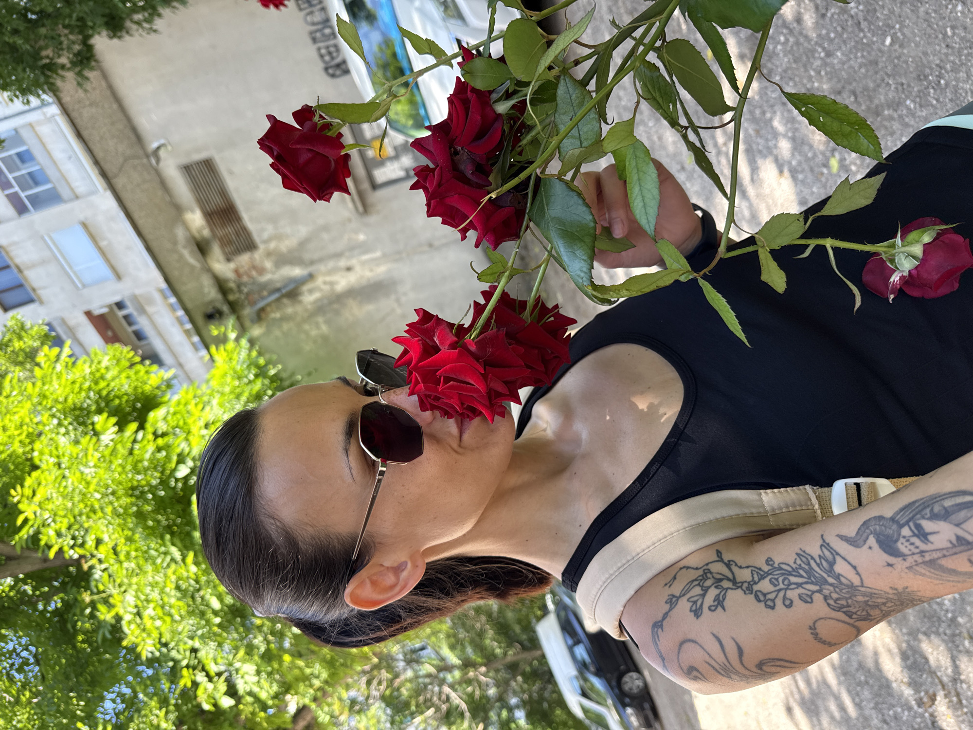 A woman with dark hair in a ponytail, wearing sunglasses and a black tank top, is holding a bouquet of red roses and smelling them. She has tattoos on her left arm. She is standing in front of a light-colored building in the city of Pleven, Bulgaria. Green trees are visible in the background, along with parked cars and some graffiti on the building wall. The sun is shining, casting shadows on the ground.