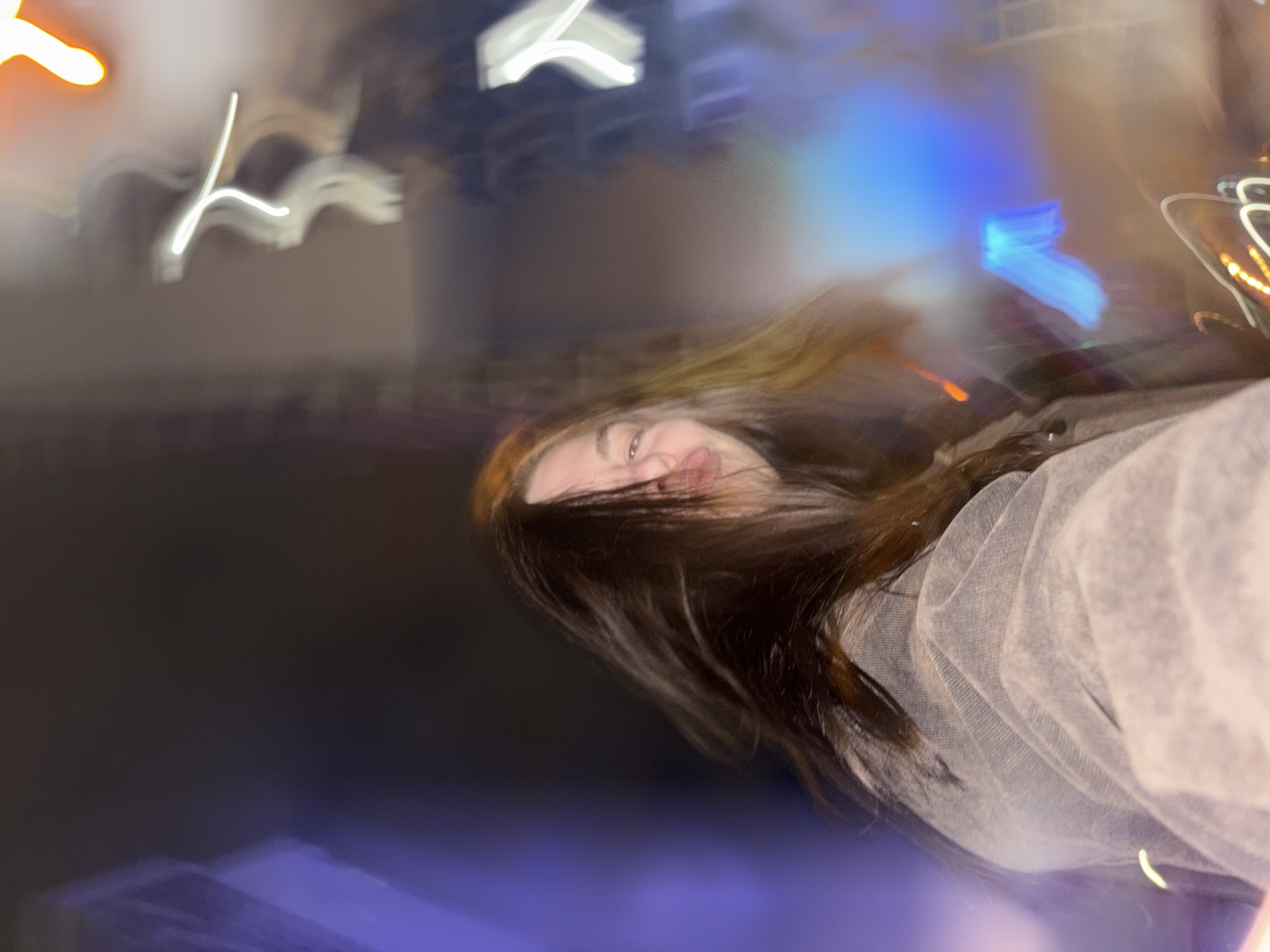 This is a blurry, long-exposure photograph taken at night in an urban environment, likely in Perm, Russia. The primary subject is a young woman, positioned diagonally in the frame. Her face is partially visible, mostly from the nose upwards, with her eyes looking towards the viewer. Her lips are slightly puckered, indicating a playful expression. Her long, dark hair is streaked and blurred due to motion, flowing downwards and across the frame. She appears to be wearing a light-colored, possibly sheer or textured fabric on her right side.
The background is dark, characteristic of nighttime. It is filled with vibrant, abstract light trails and blurred light sources. Prominent light elements include elongated white streaks and amorphous shapes in the upper left and central top portions, a distinct bright blue rectangular light on the mid-right, and accents of orange, yellow, and purple light scattered throughout. These lights suggest the presence of city illumination, such as building lights or neon signs.
The overall impression is one of movement and a dynamic urban night scene, possibly captured while walking or in transit. The significant motion blur gives the image a dreamy and artistic quality. The woman's expression conveys a sense of lightheartedness or fun. There is no visible text in the image.