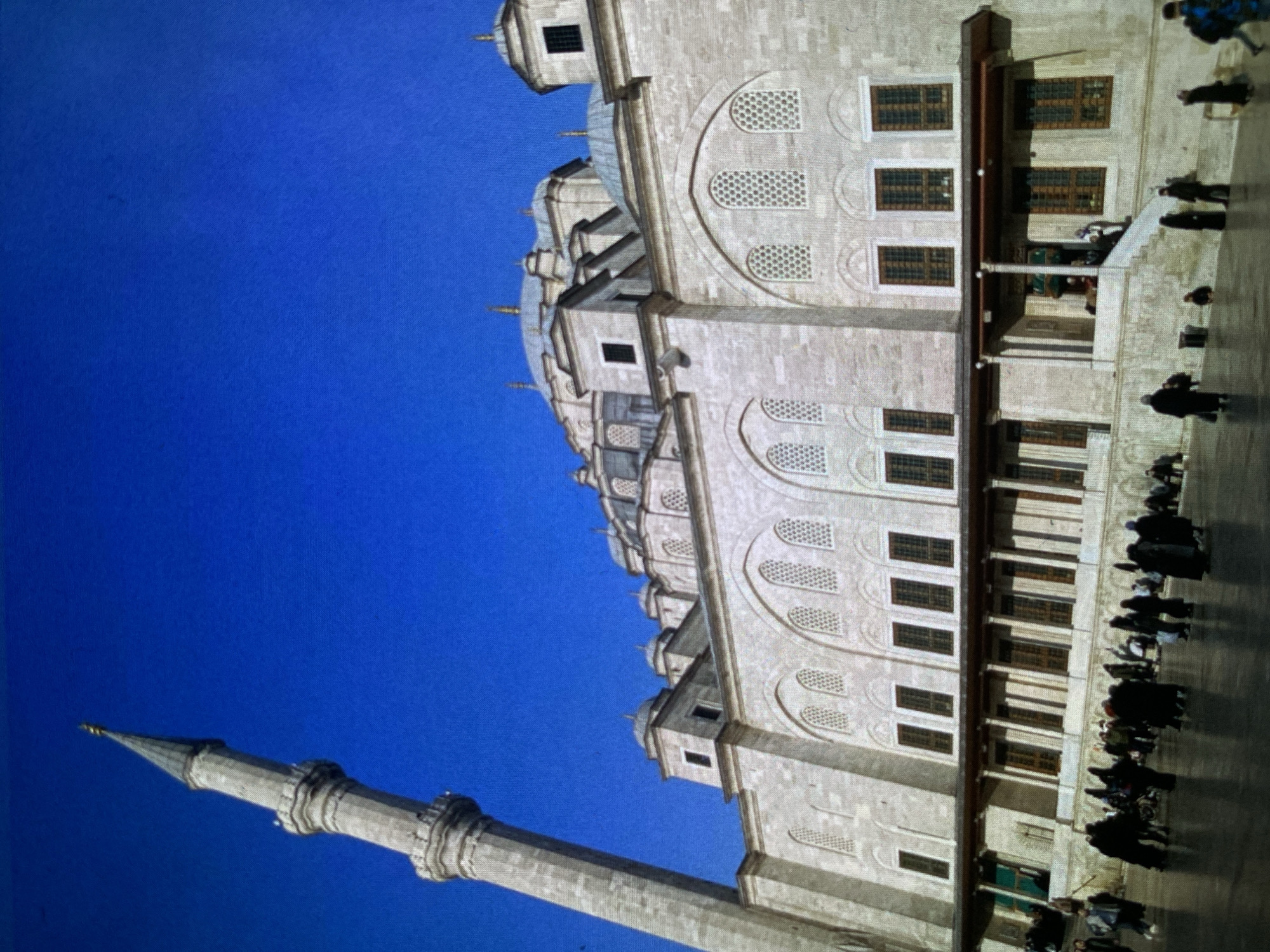 The image displays a grand, light-colored stone building, characteristic of an Ottoman imperial mosque, set against a vibrant, clear blue sky. The structure features multiple domes of varying sizes, intricate arched windows with decorative grilles, and a richly detailed facade. A slender, towering minaret, built of light stone with multiple balconies, extends upwards on the left side of the frame.
On the ground level of the building, numerous people are visible. They appear to be visitors or congregants, walking individually or in small groups, with some gathered near the building's entrance area which features columns. The activity suggests a public gathering or tour at a religious or historical site.
The clear, bright blue sky indicates good weather during the daytime. The overall scene is calm and showcases impressive architecture. No discernible text is visible. The location context provided is Bankya, Bulgaria.