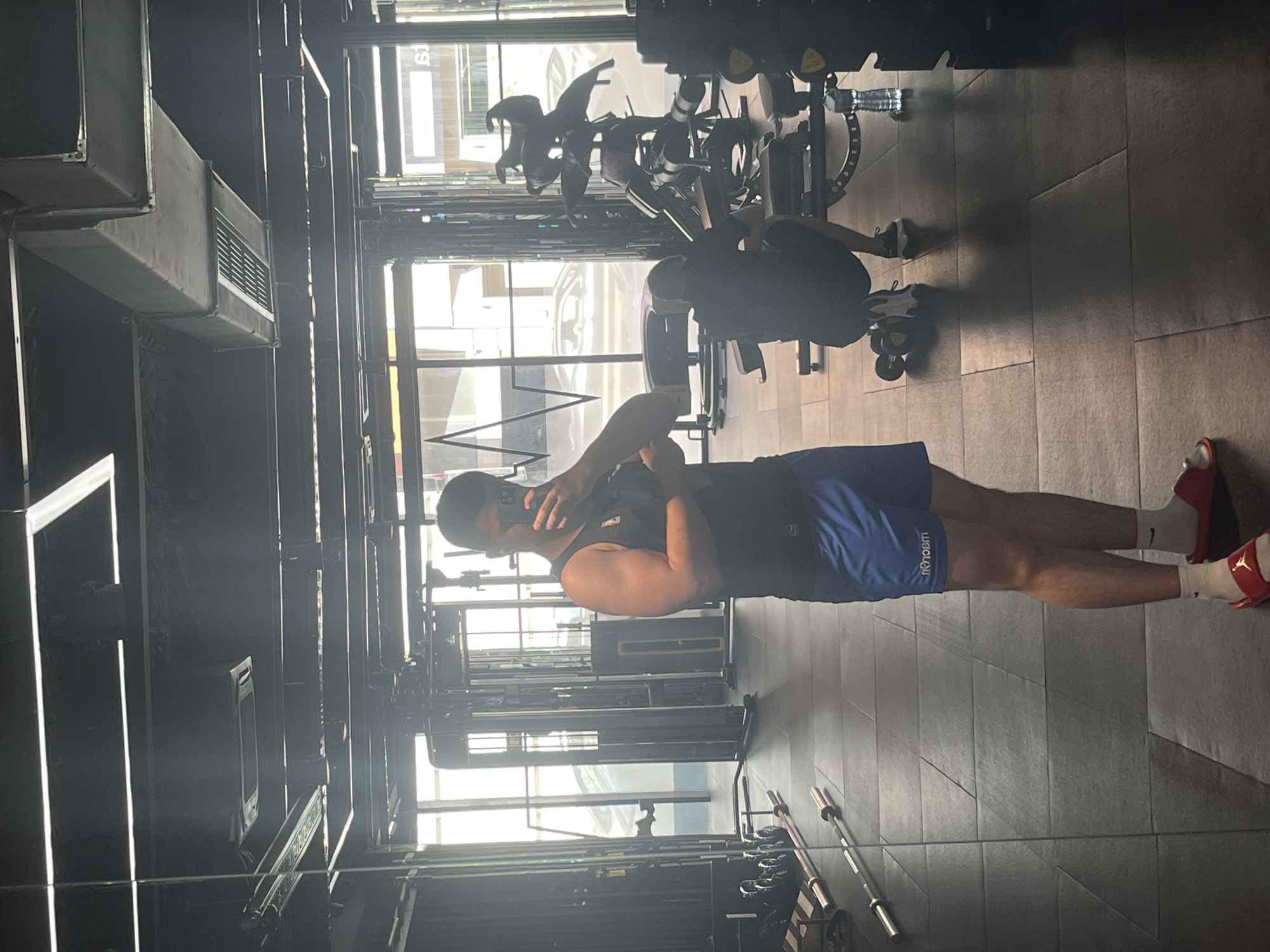 A man is captured in a mirror selfie within a brightly lit, modern gym. He stands upright, wearing a black tank top, blue shorts with the reversed text "UBCLAU" (likely "LUCLAU" when read correctly), white Nike socks, and red Jordan brand slides featuring the Jumpman logo. He holds a phone with his right hand, obscured by it, to take the picture.
The gym features dark walls and a tiled floor, creating a sleek, industrial aesthetic. Large windows dominate one side, allowing ample natural light to flood the space and create reflections of the exterior, including indistinct cars and buildings. Various gym equipment is visible, such as barbells, weights, and exercise machines. In the background, another person is partially visible near some equipment. Exposed air ducts are prominent on the ceiling. The presence of bright natural light indicates it is daytime. A water bottle is also seen on a piece of equipment.