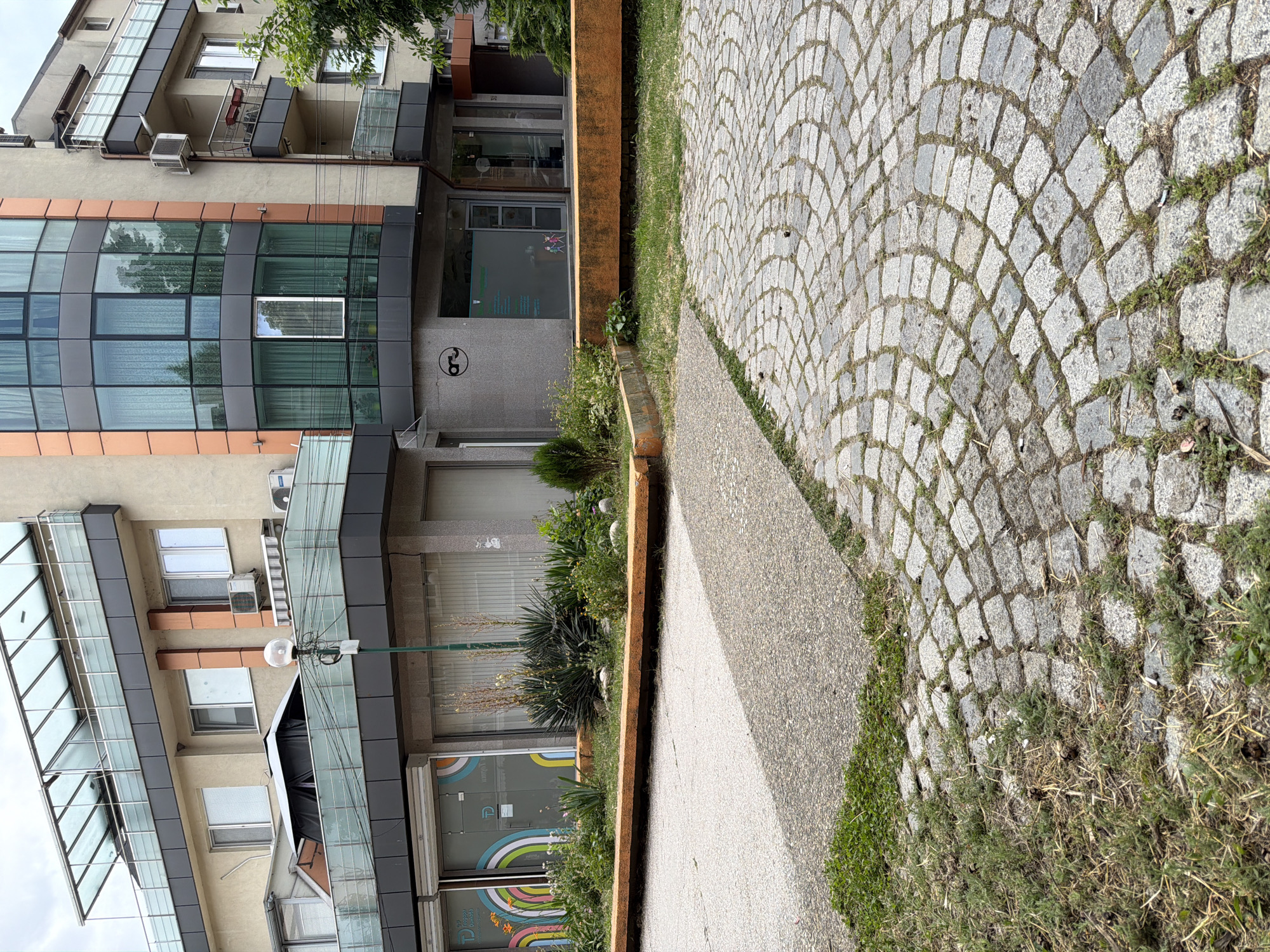 The image captures a street-level view of a modern building facade in Pazardzhik, Bulgaria, characterized by its contemporary architecture. The building features a multi-story design with a prominent glass-enclosed corner, an orange-colored accent on the facade, and balconies with glass railings. Below the main structure, there are commercial storefronts with large windows, one of which displays colorful abstract art. The ground level is a mix of paved surfaces: a section of asphalt and a circular pattern of cobblestones. Greenery, including grass and plants, is visible in planters and along the edges of the pathways. A street lamp stands to the left of the frame, and outdoor seating elements, possibly benches, are integrated into the landscaping in front of the storefronts. The sky appears overcast.