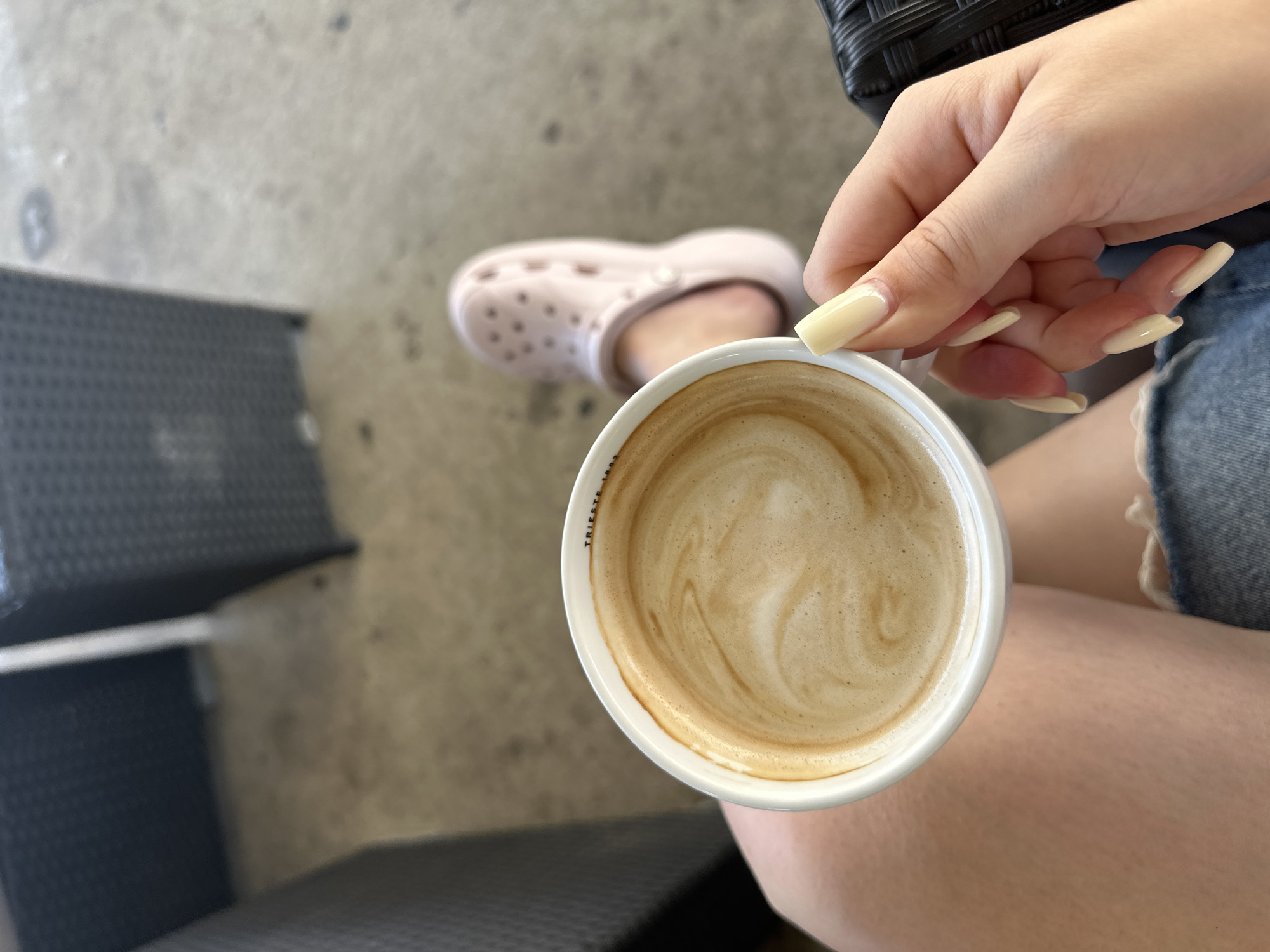 A close-up shot captures a person's hand holding a white mug of coffee with latte art. The person's legs, clad in denim shorts, are visible in the foreground. In the background, a light pink Croc shoe is on a foot, suggesting an outdoor seating area with a textured surface. The scene appears to be an outdoor cafe, possibly in the city of Grad Rijeka, Croatia, judging by the style of outdoor furniture and pavement. The overall impression is one of relaxed enjoyment of a beverage in a public setting.