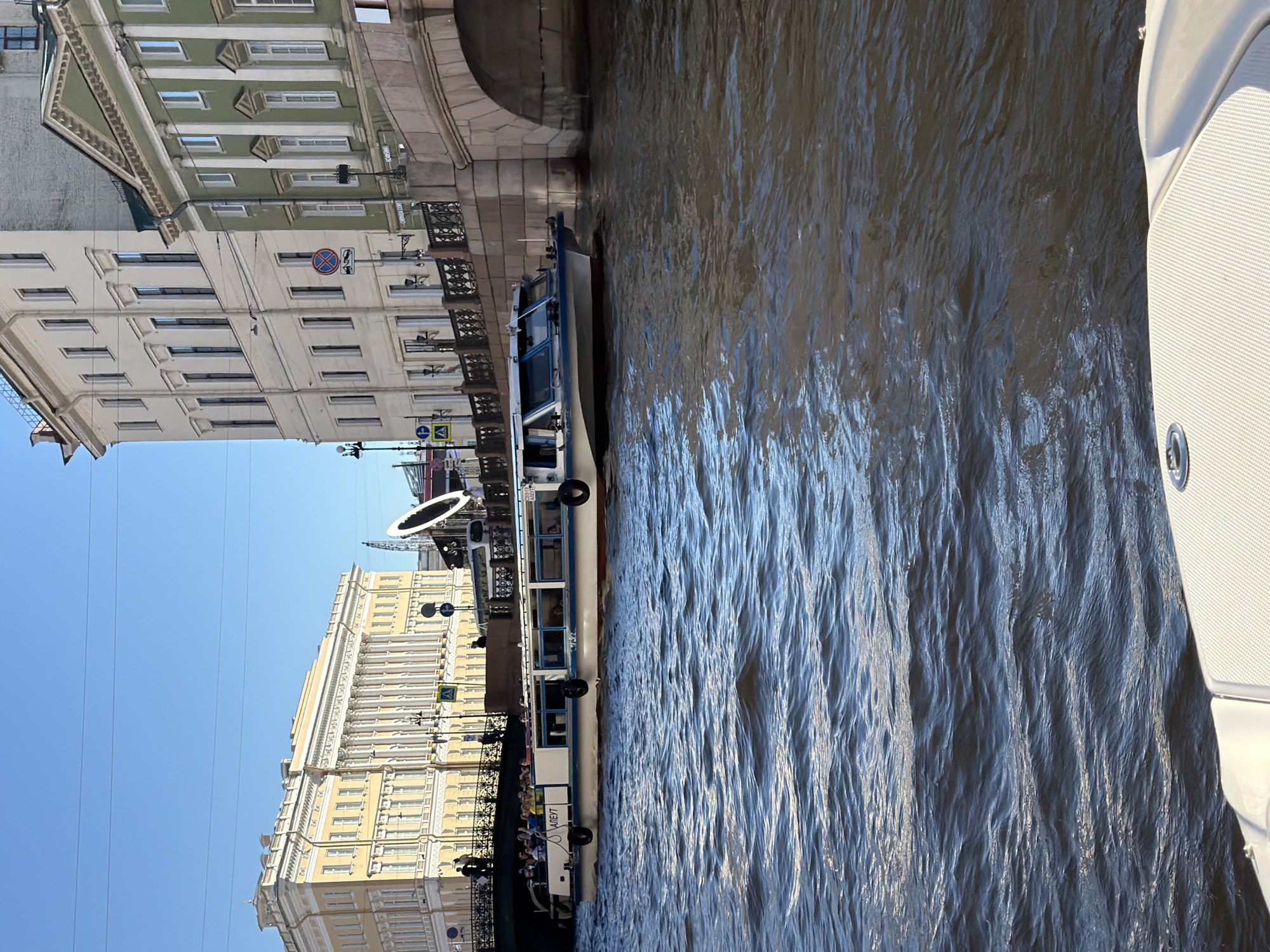 A tour boat full of passengers navigates a river in Saint Petersburg, Russia, under a clear blue sky. The boat, which has "АЛБУТ" written on its side, is white with a blue stripe and features several windows. It is passing by a bridge and several ornate buildings. The architecture suggests a historical district, with the buildings featuring classical elements such as columns and arched windows. The river is choppy, with sunlight reflecting off the water's surface. The foreground shows the edge of another boat, also white and seemingly part of the waterway's infrastructure.